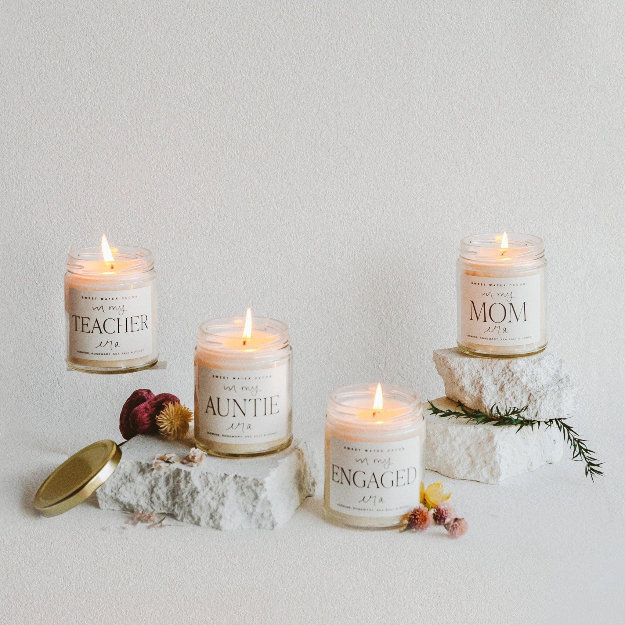 Four personalized soy candles with gold lids arranged on white sand with dried flowers and a gold spoon, labeled Teacher, Auntie, Engaged, and Mom.
