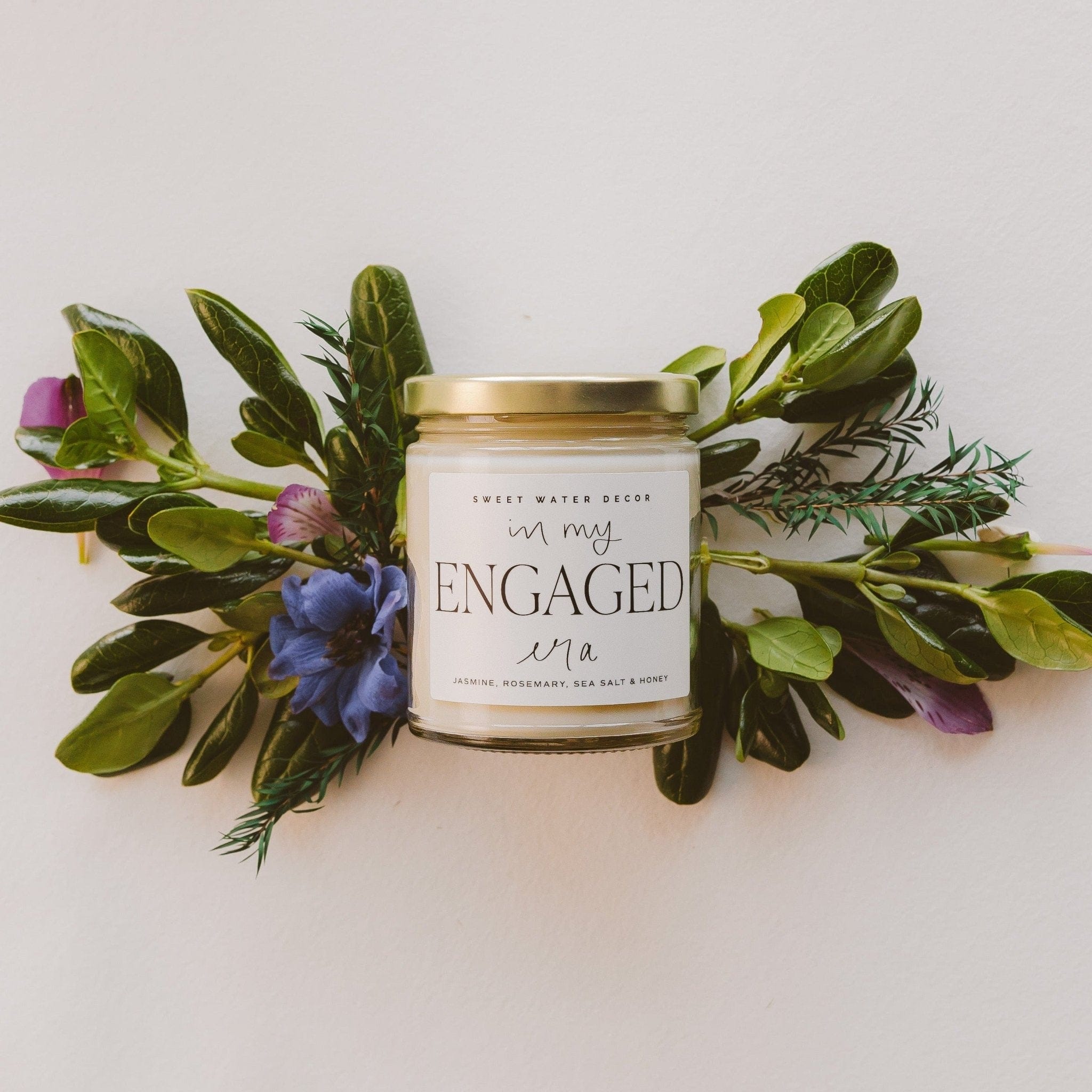 White soy candle jar with gold lid surrounded by fresh green foliage and purple wildflowers, labeled "In My Engaged Era"