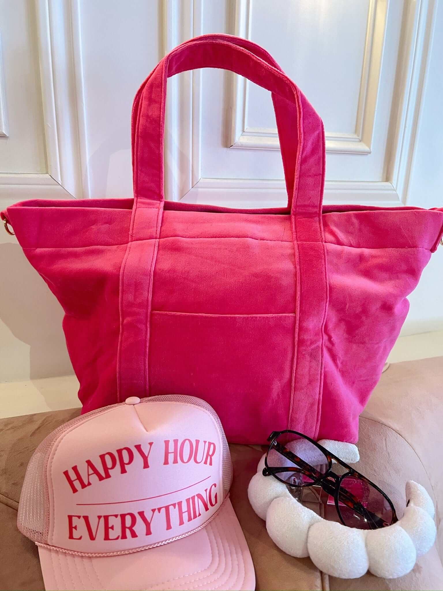 Bright pink canvas tote bag with matching pink trucker hat reading "Happy Hour Everything" and fuzzy white slippers displayed on wood floor.