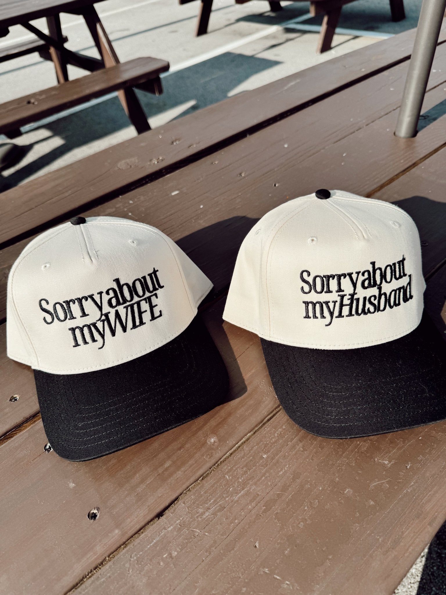 Two vintage trucker hats with white and black colorway sitting on wooden deck, one reading "Sorry about my Wife" and the other "Sorry about my Husband"