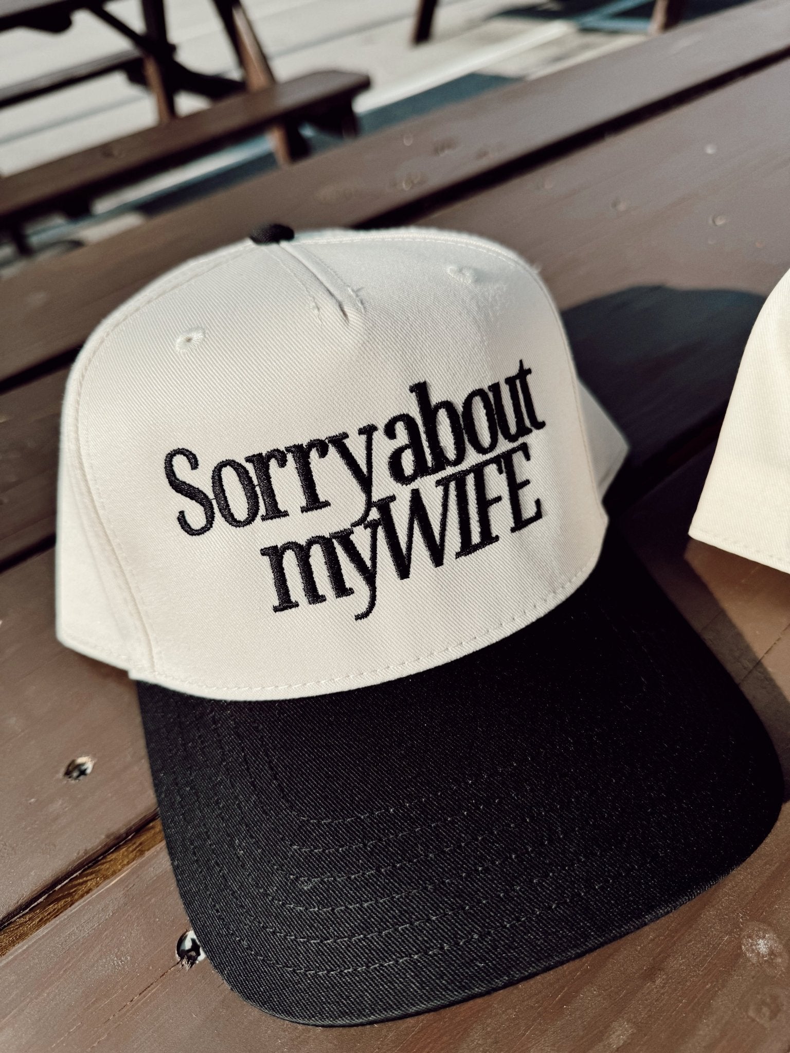 White and black baseball cap with "Sorry about my WIFE" text printed on the front panel, sitting on a wooden surface.