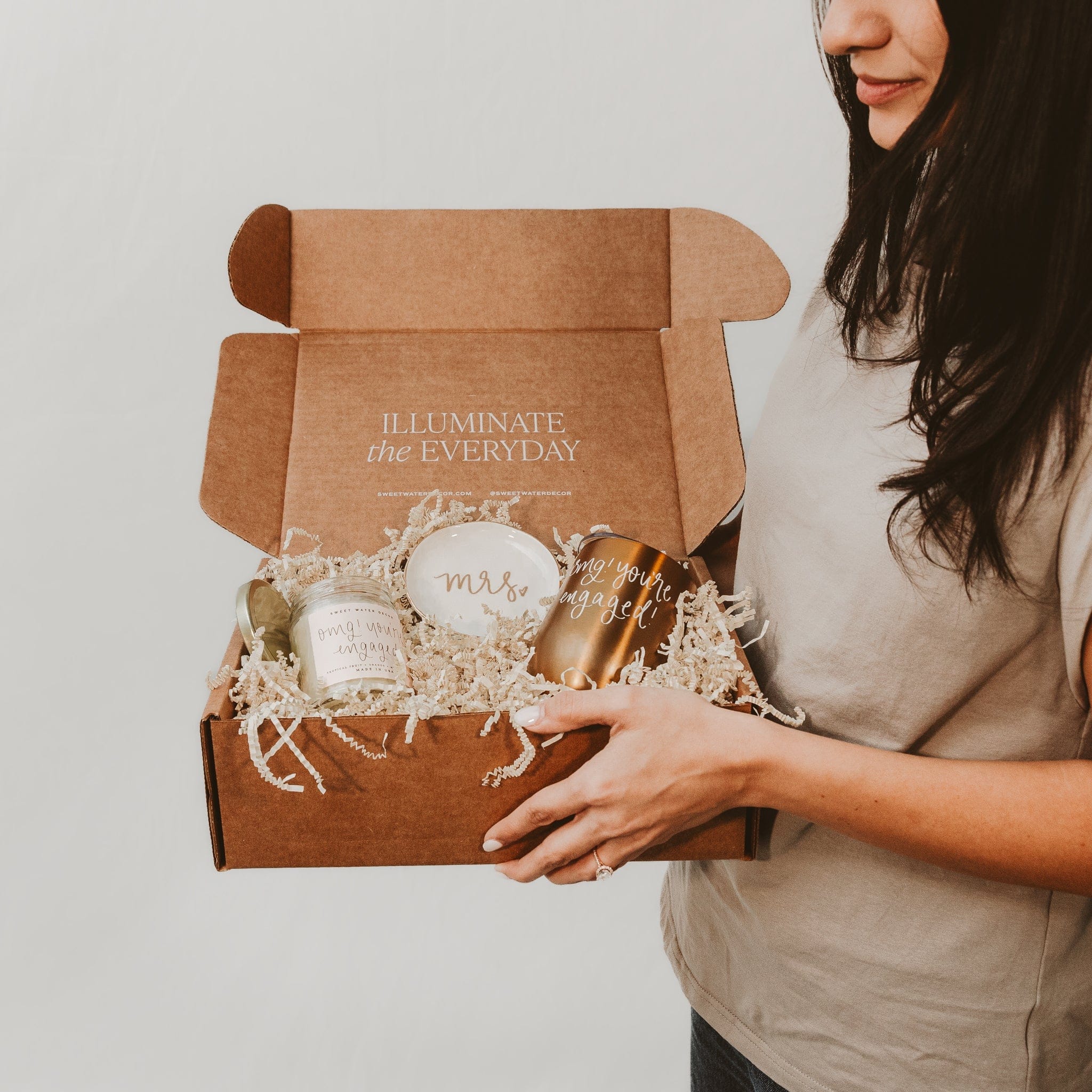 Woman holding open kraft gift box filled with candle, soap, and gold accessories with "Illuminate Her Everyday" text.