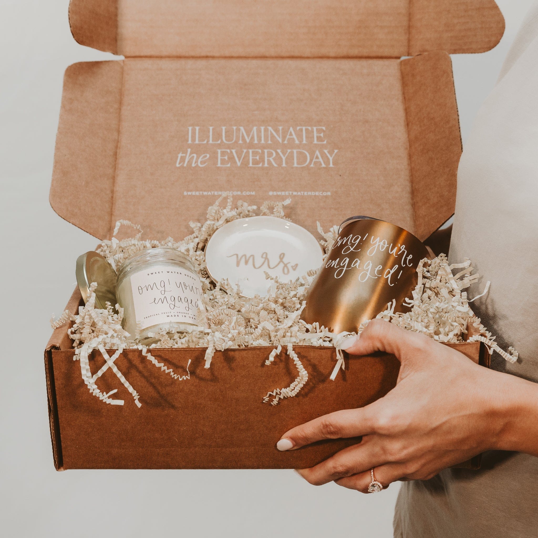 Kraft gift box with "ILLUMINATE the EVERYDAY" text, containing candles, a gold bottle, and white ceramic vessels nestled in cream tissue paper.