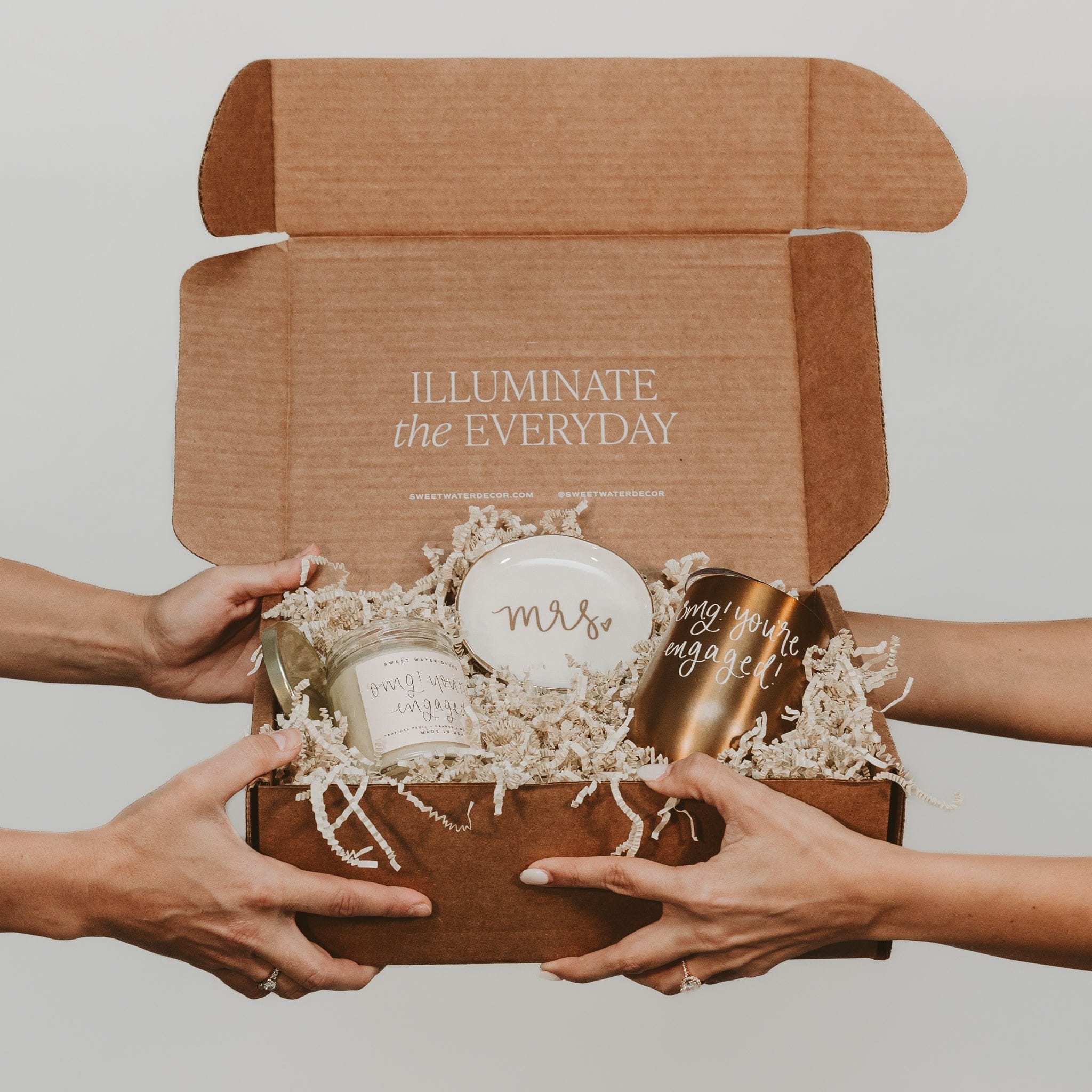 Brown kraft gift box with "Illuminate the Everyday" text, held by two people's hands, containing personalized Mrs. mug and gold accessories nestled in white shredded paper.