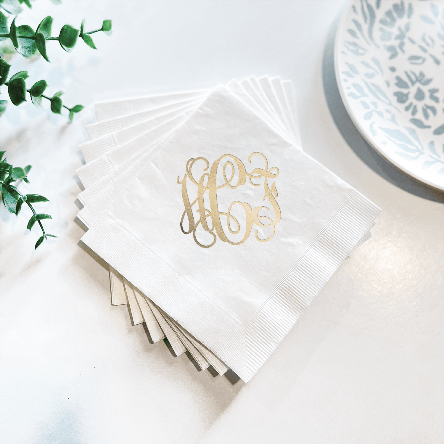 Stack of white napkins with gold foil monogram design, styled with blue patterned plate and greenery