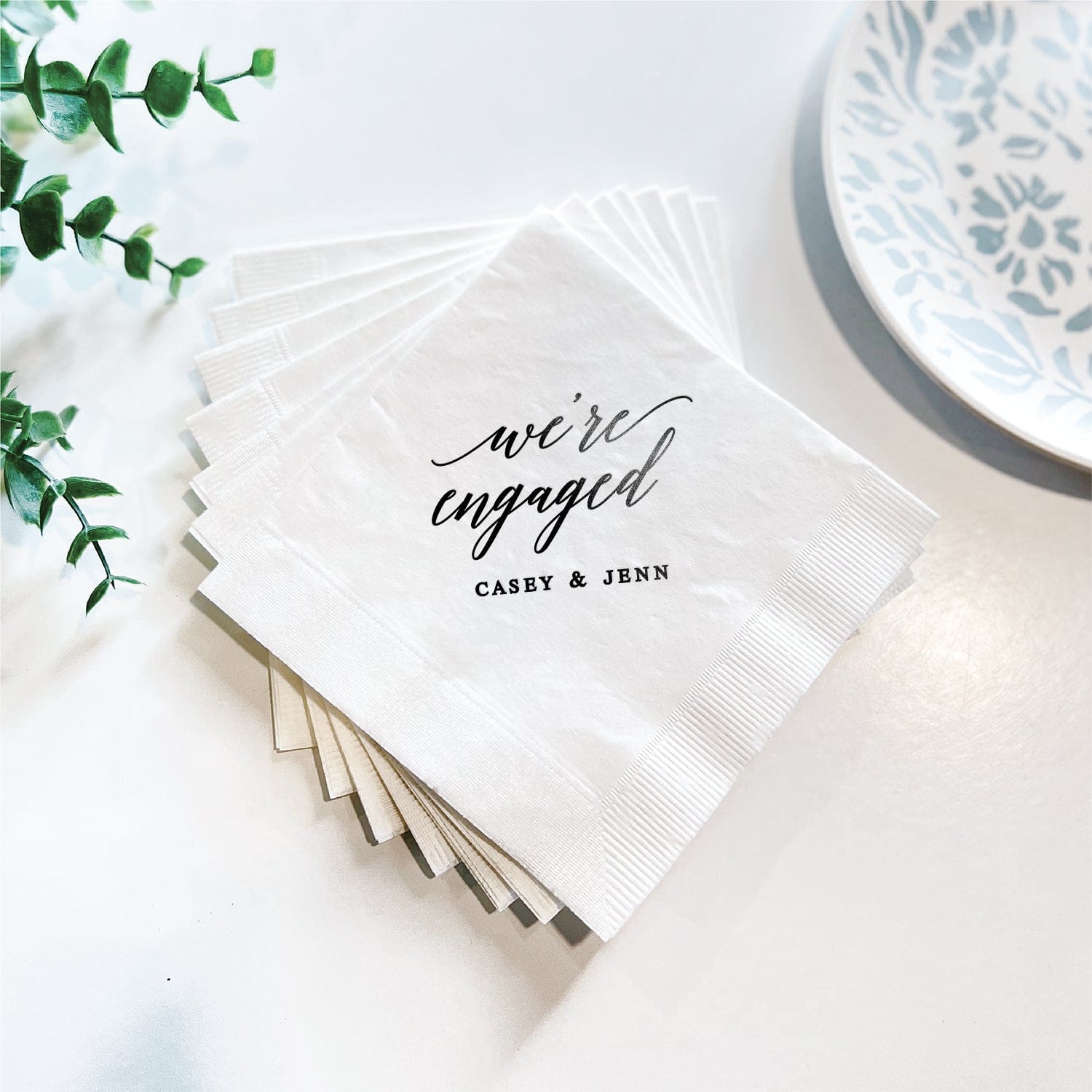 White foil-stamped napkins with "we're engaged" script and couple names Carey & Jenn printed in black and silver ink.
