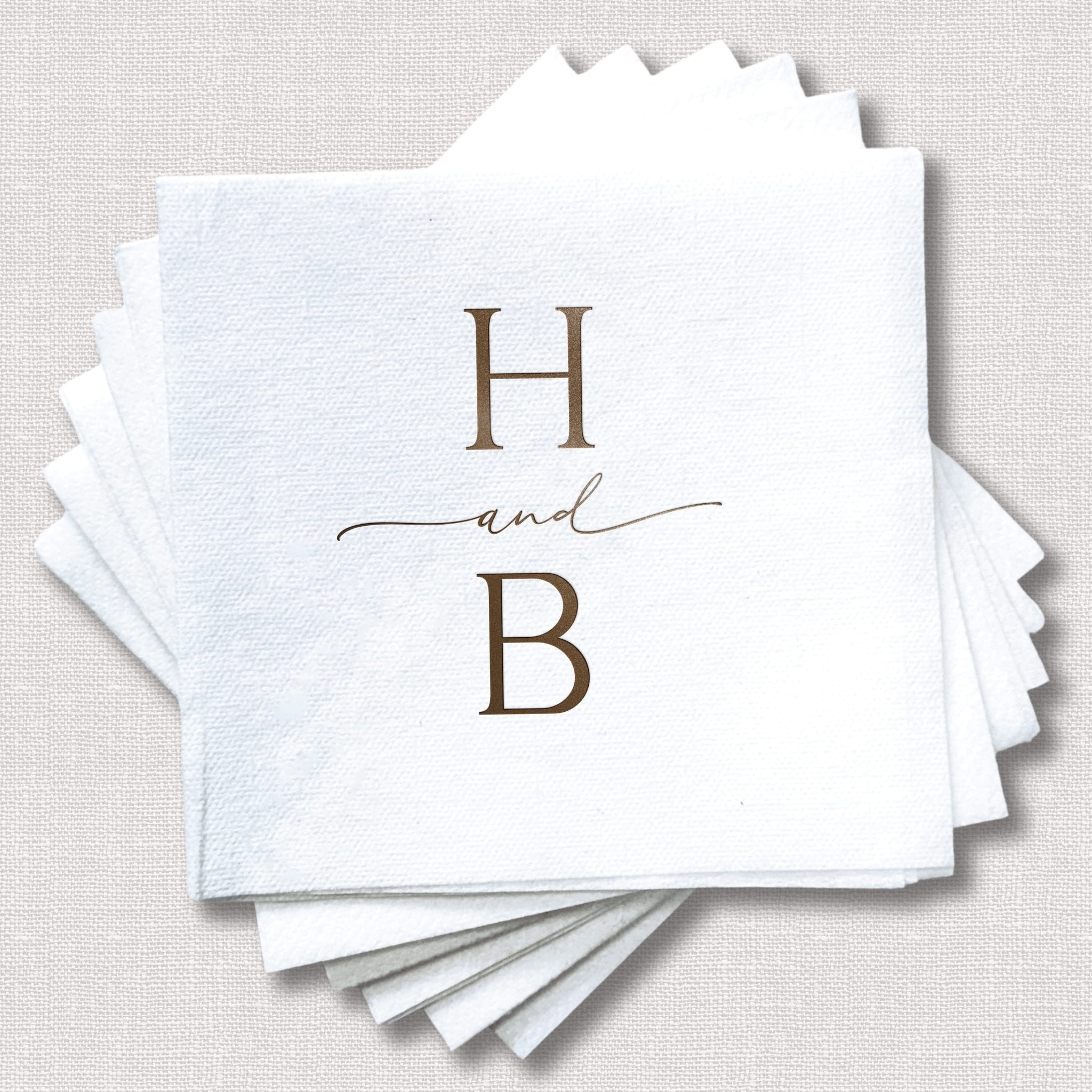 Stack of white linen cocktail napkins with gold foil stamped initials H and B with decorative line detail