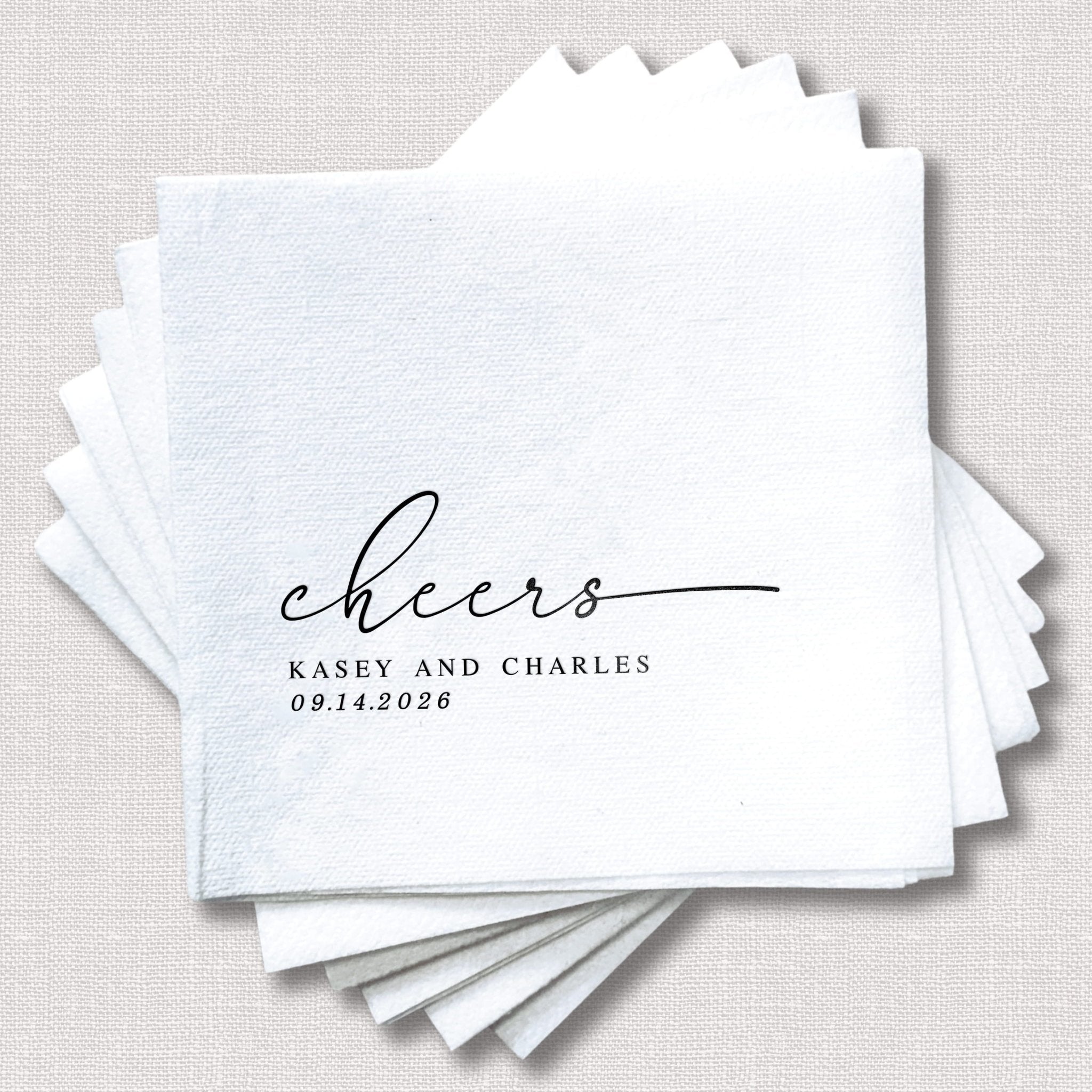 Stack of white linen napkins with black script text reading "cheers" and "Kasey and Charles 09.14.2028" personalized for a wedding.