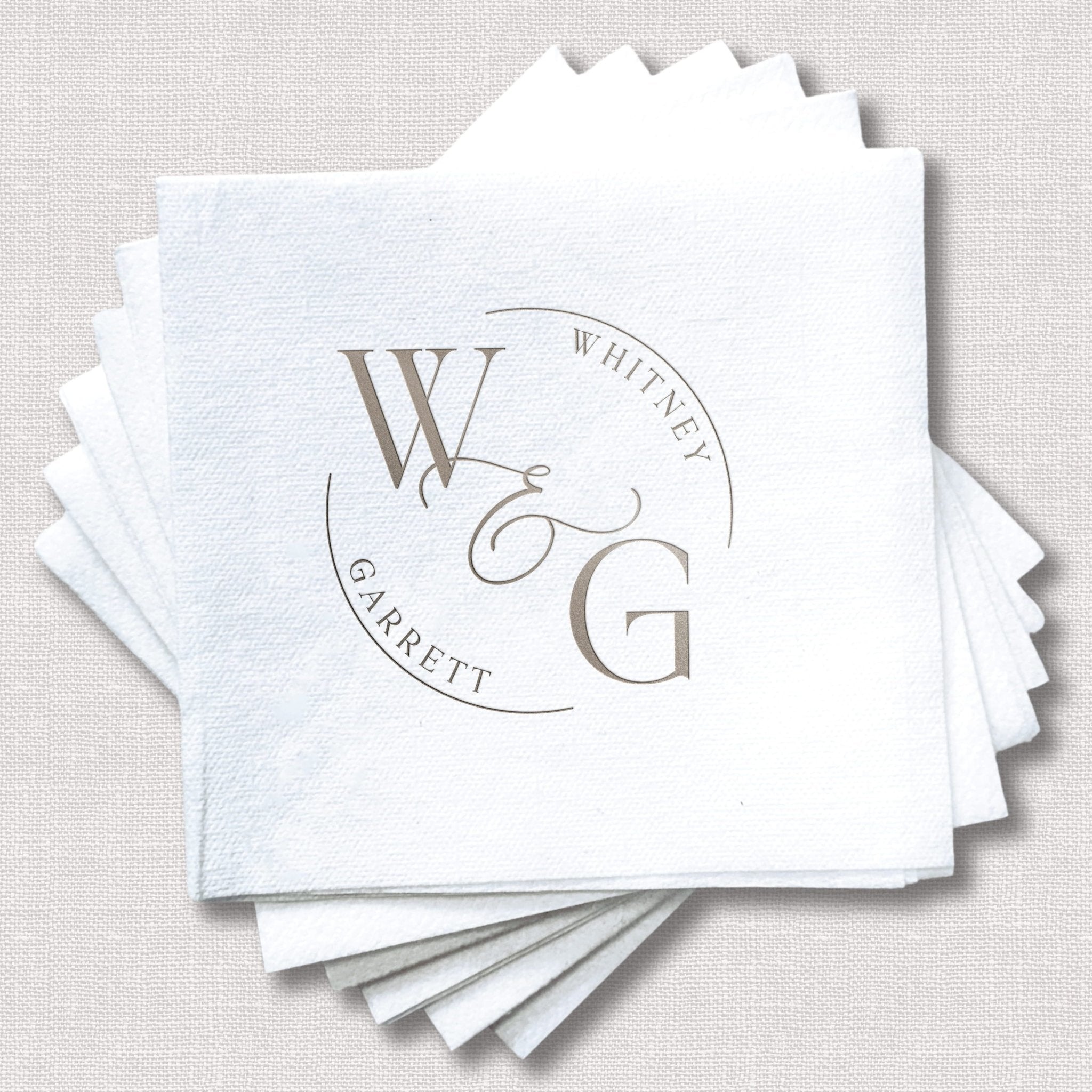 Stack of white linen cocktail napkins with gray foil stamped monogram and "WHITING GARRETT" wedding design in circular layout.