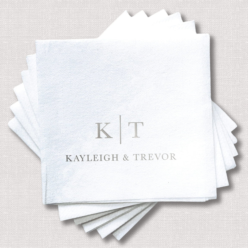Stack of white linen napkins with gray foil-stamped initials K|T and names Kayleigh & Trevor for a wedding.