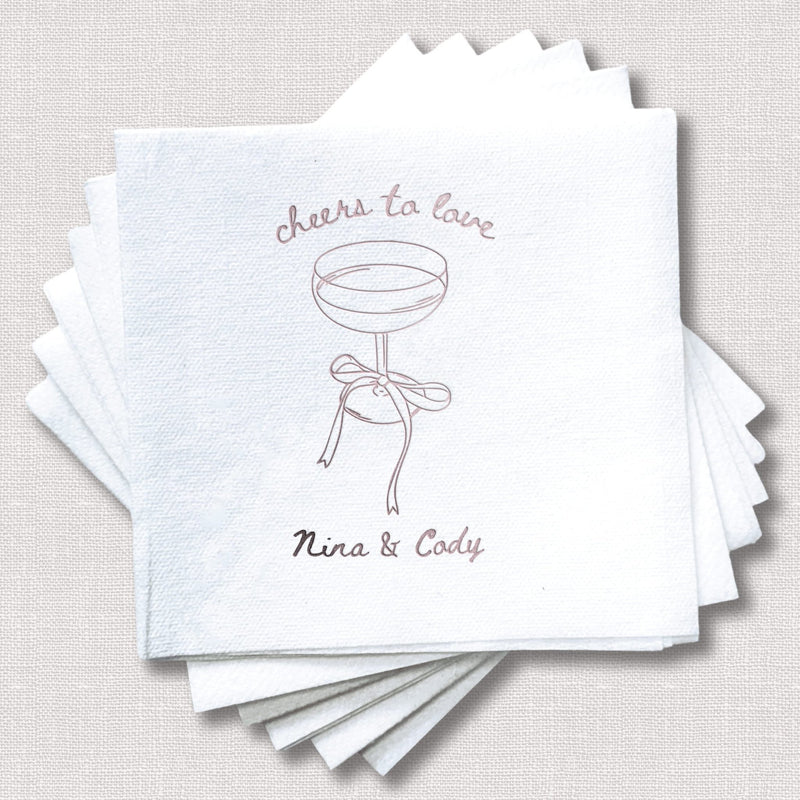 Stack of white linen napkins with "cheers to love" text, cocktail glass illustration, and personalized names "Nina & Cody" in rose gold foil.