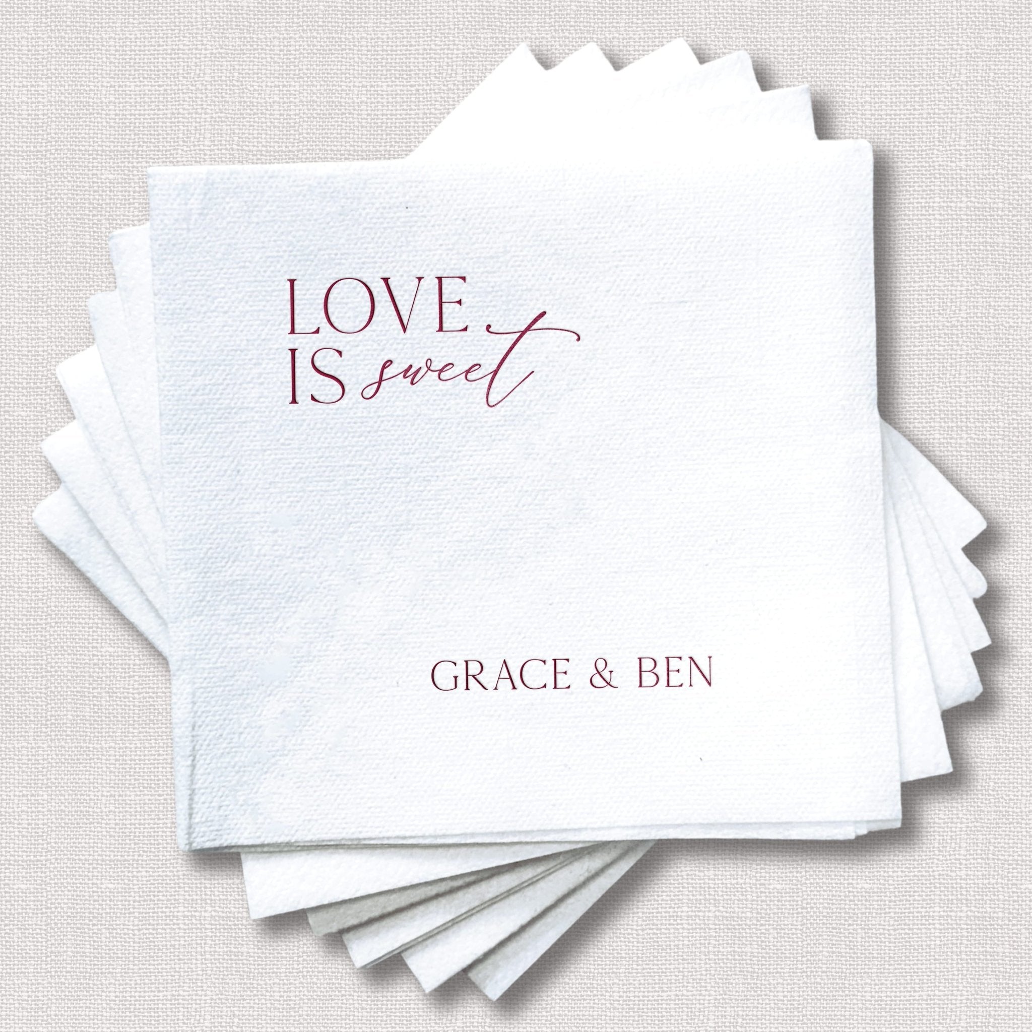 Stack of white linen napkins with scalloped edges, featuring "Love Is Sweet" text in burgundy script and personalized names "Grace & Ben" in gray.