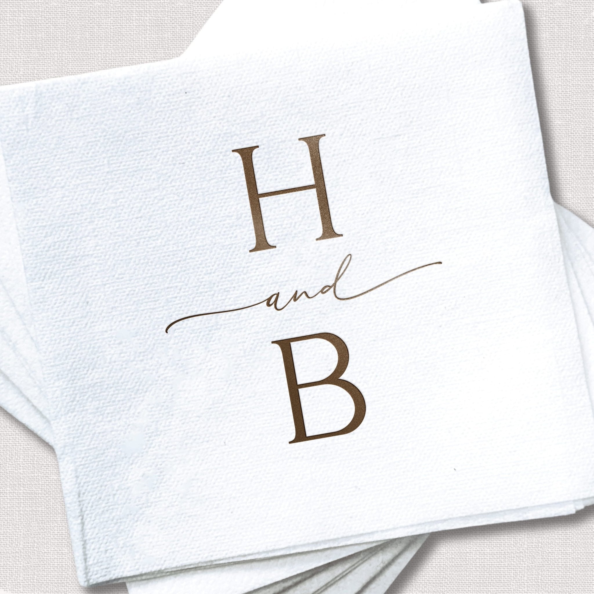 White linen cocktail napkins with brown foil stamped initials H and B connected by an ampersand design