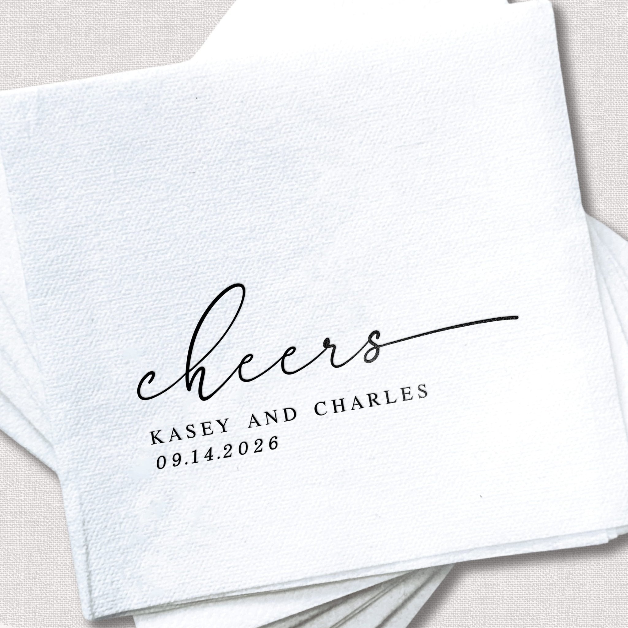 White linen napkins personalized with "cheers" script and "Kasey and Charles 09.14.2026" in black foil stamping.