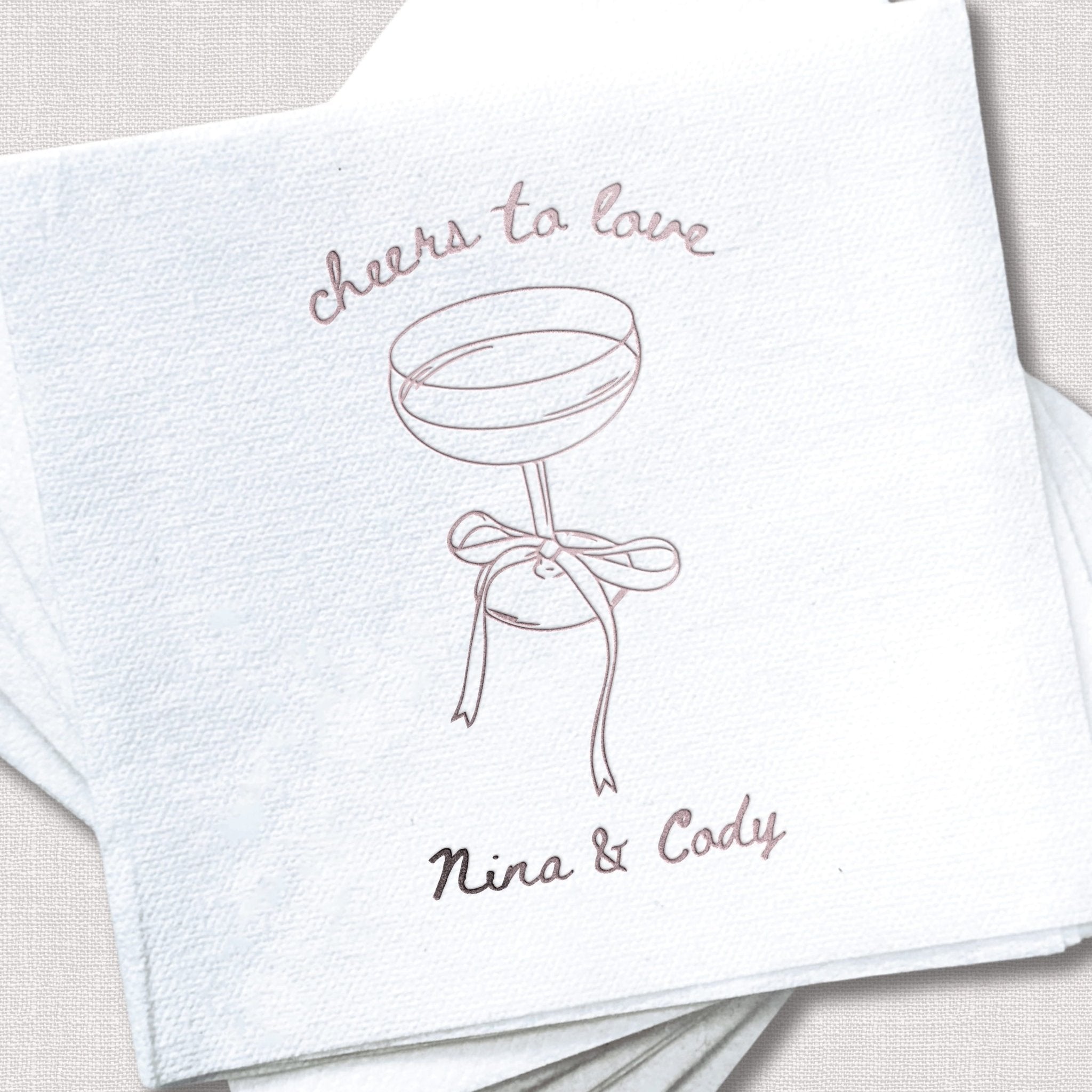 White linen napkin with "cheers to love" text, cocktail glass illustration, and couple's names Nina & Cody in rose gold foil stamping.