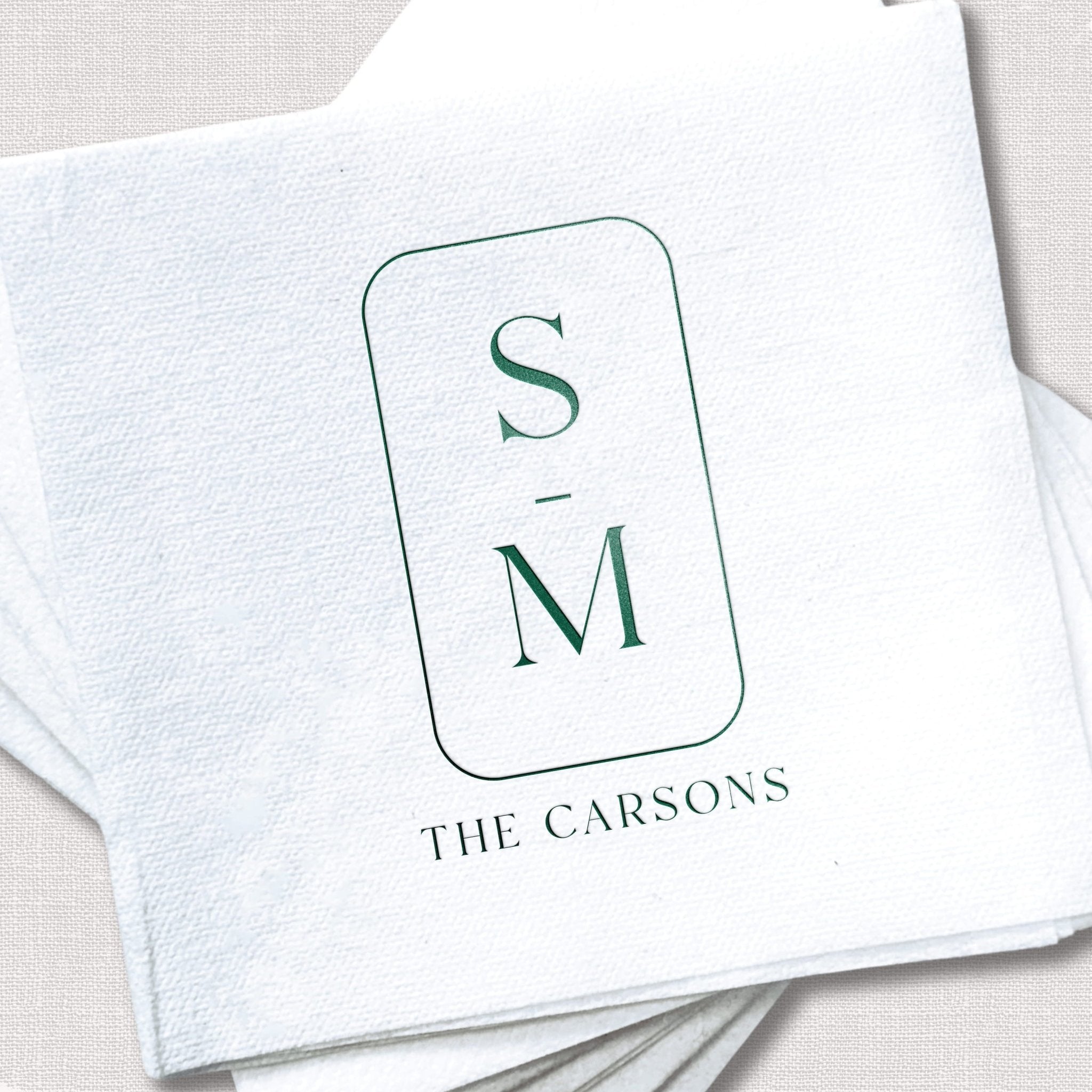 White linen napkin with green foil-stamped initials S and M in a rounded rectangle, text reading "THE CARSONS" below