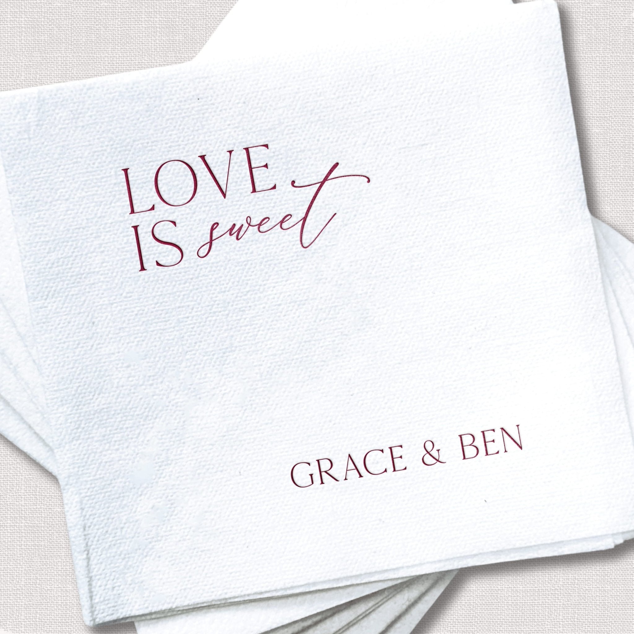 White linen napkin with rose gold foil stamped text reading "LOVE IS SWEET" and personalized names "GRACE & BEN" below.