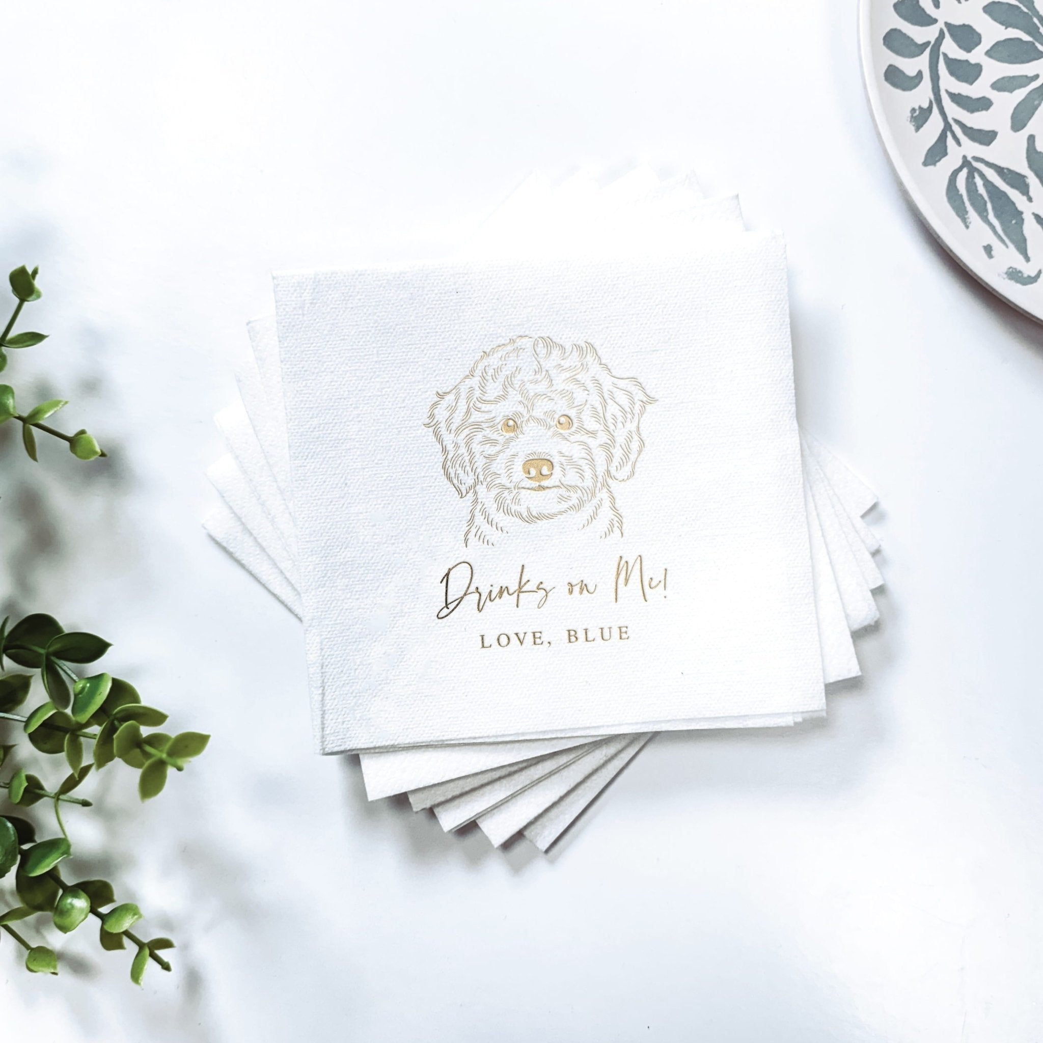 Stack of white linen napkins with gold foil stamped dog illustration and "Drinks in Paw, Love, Blue" text.