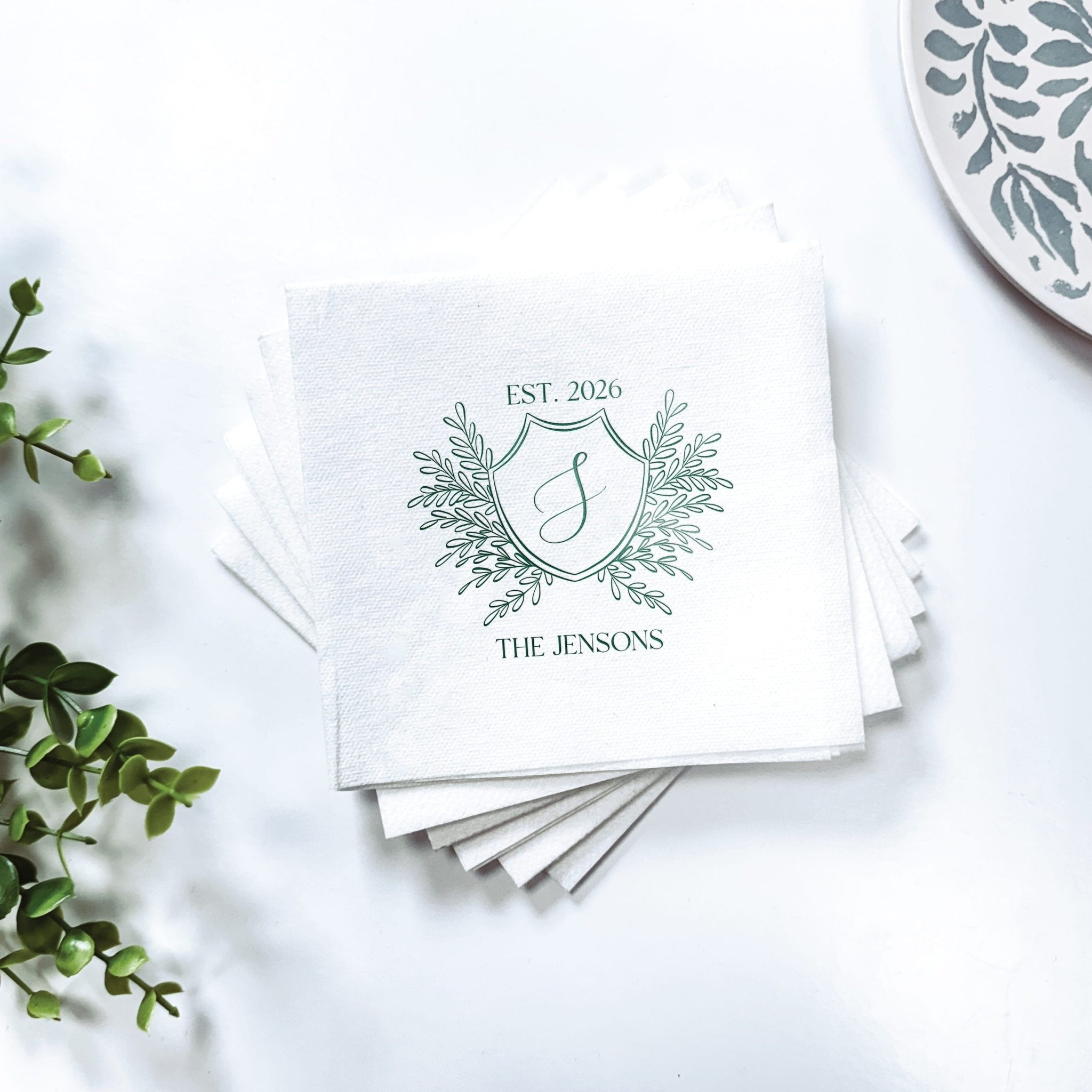 Stack of white linen napkins with custom family crest design featuring EST. 2020 and "The Jensons" text with foil stamped details.