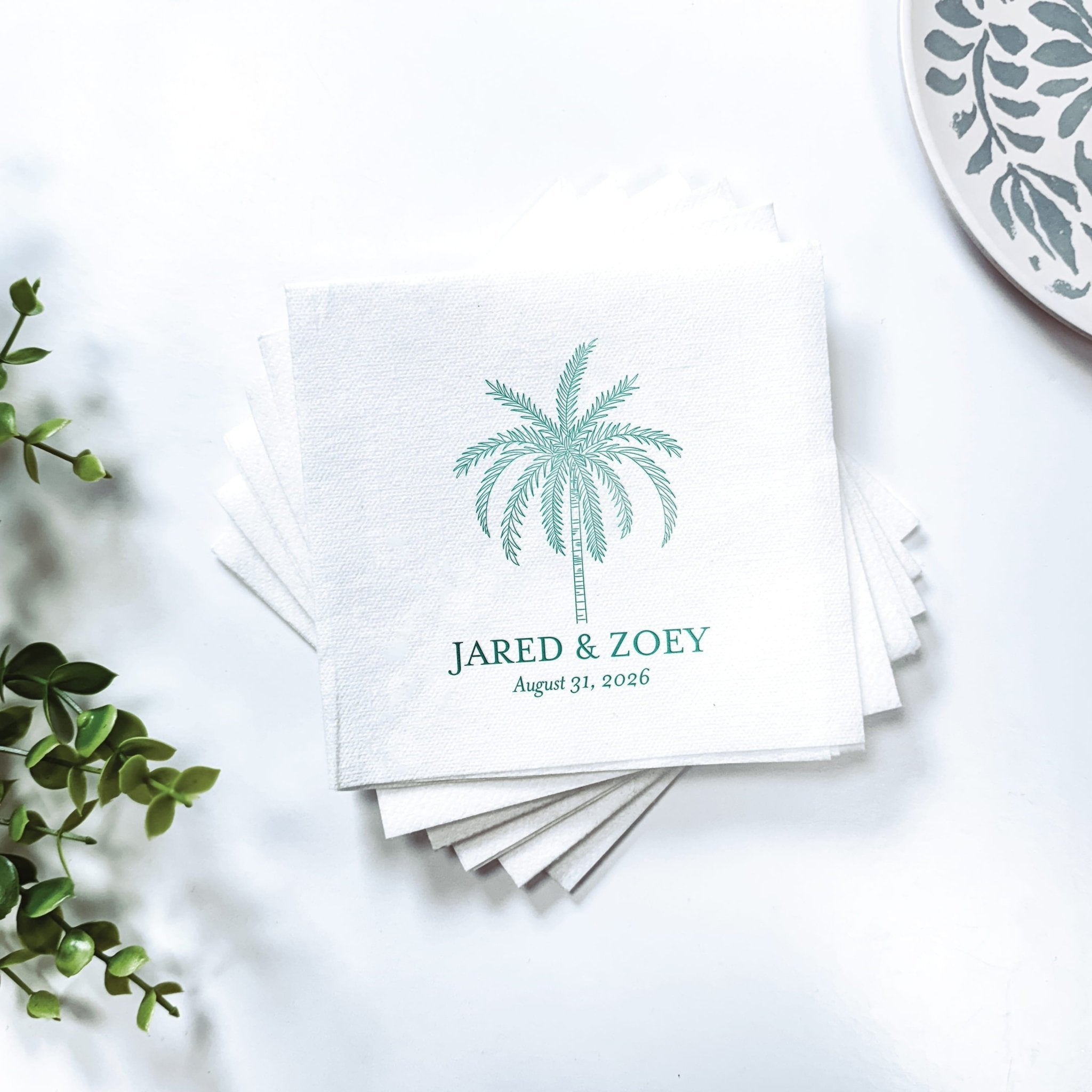 Stack of white napkins with teal palm tree design and personalized names "Jared & Zoey" with wedding date printed below