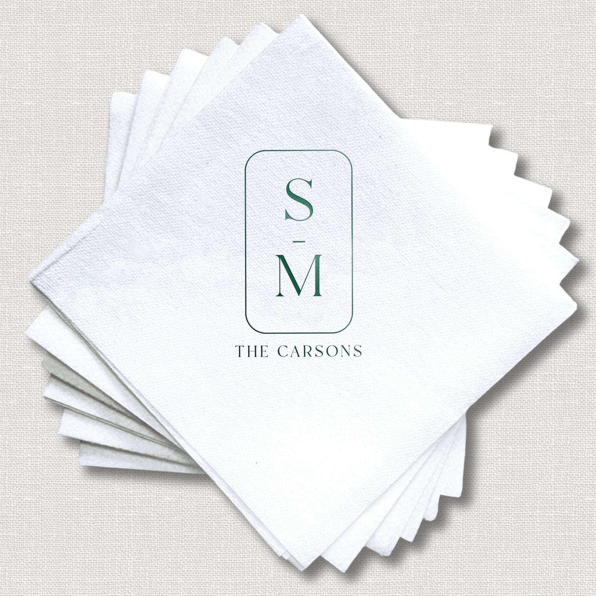 Stack of white linen napkins with green foil-stamped initials S and M and text "The Carsons" in elegant design