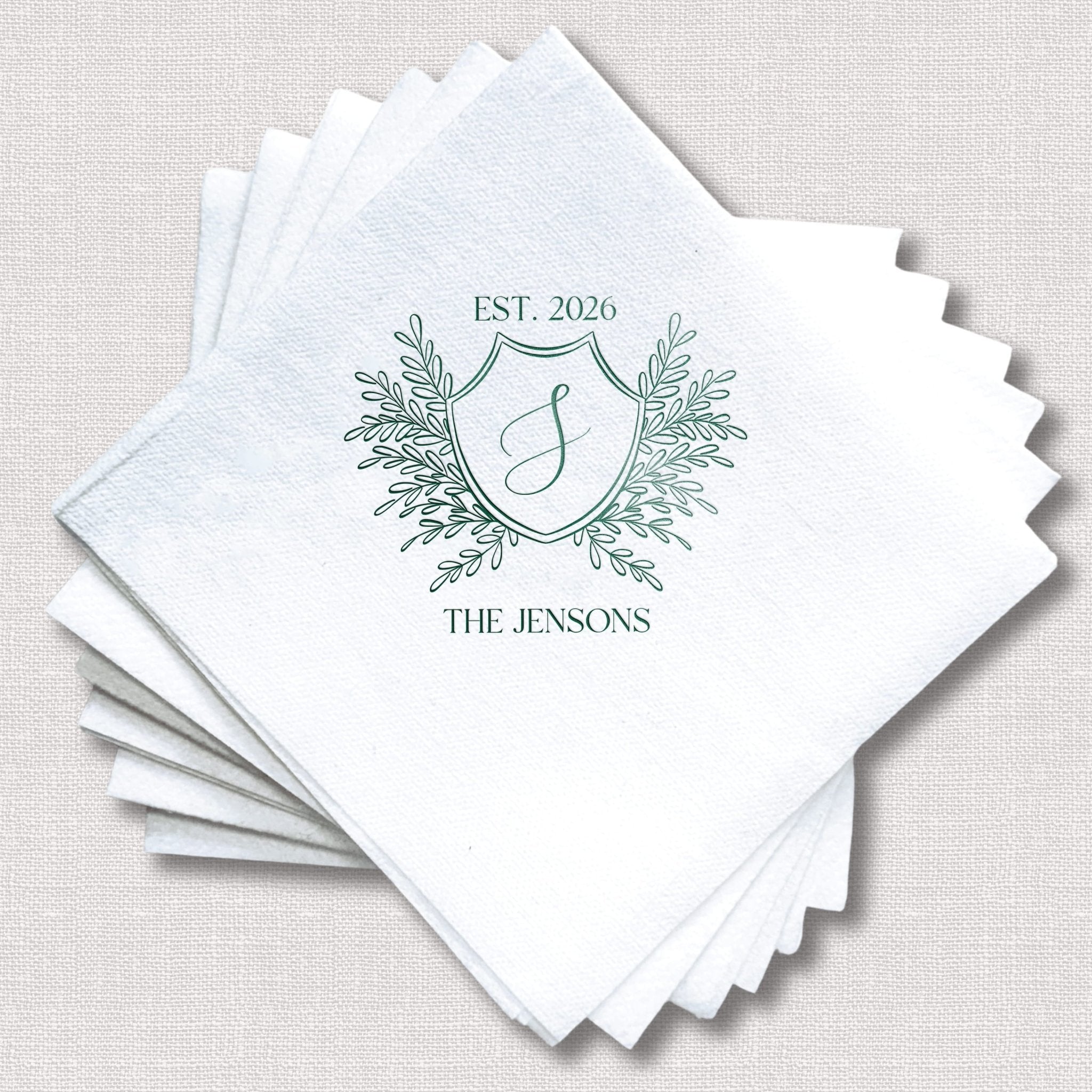 Stack of white linen napkins with green foil-stamped crest design featuring "EST 2026" and "THE JENSONS" personalization.