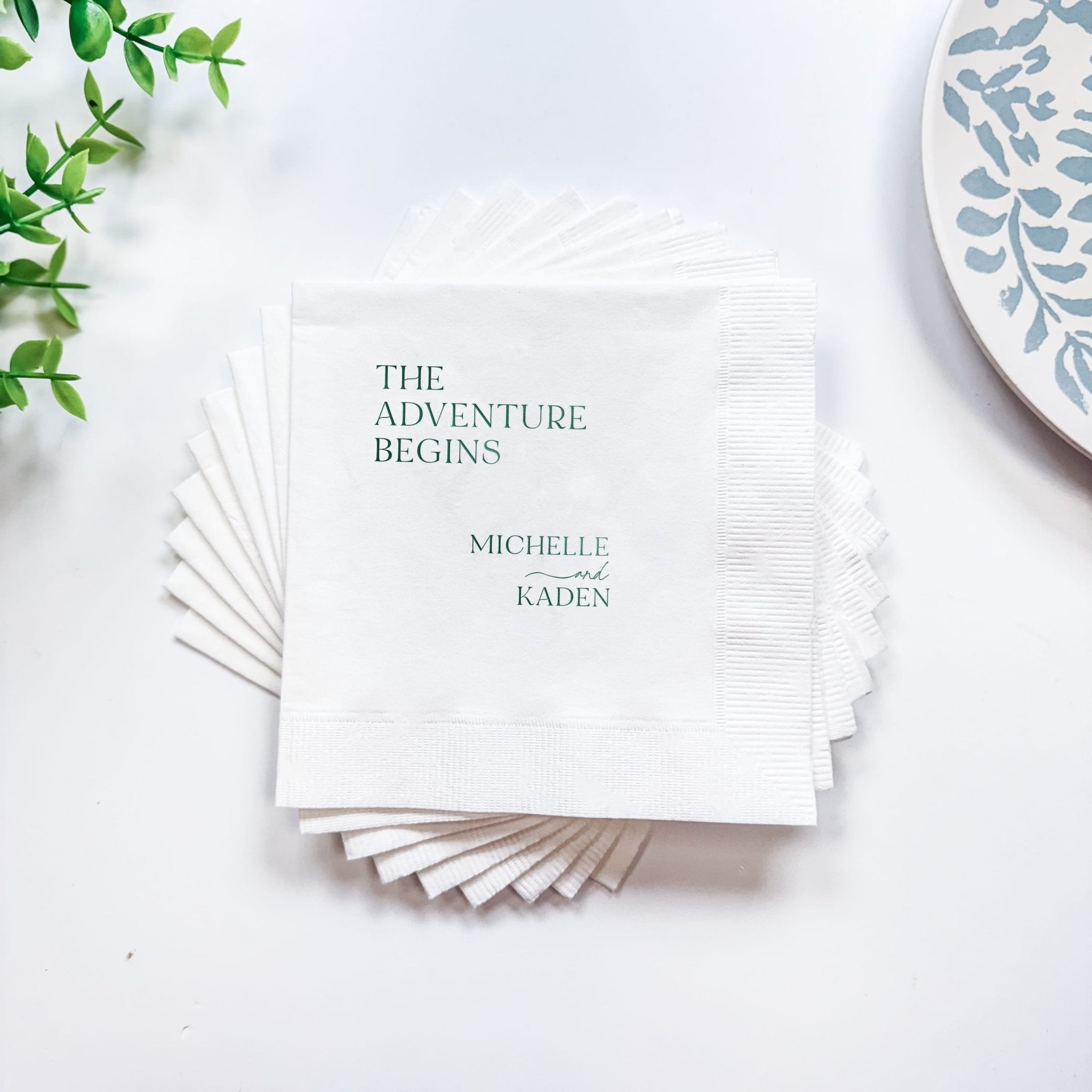 Stack of white foil stamped napkins with "The Adventure Begins" and personalized names Michelle and Kaden printed in gray text