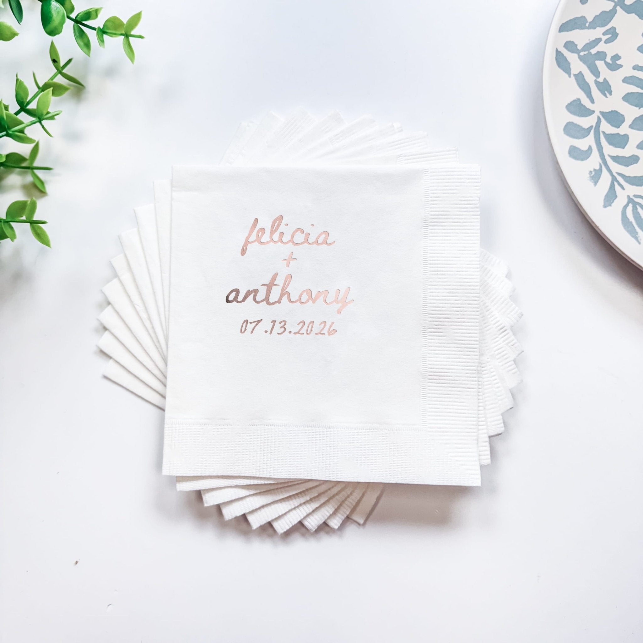 Stack of white cocktail napkins with rose gold foil-stamped names "alicia anthony" and date "07.13.2026"