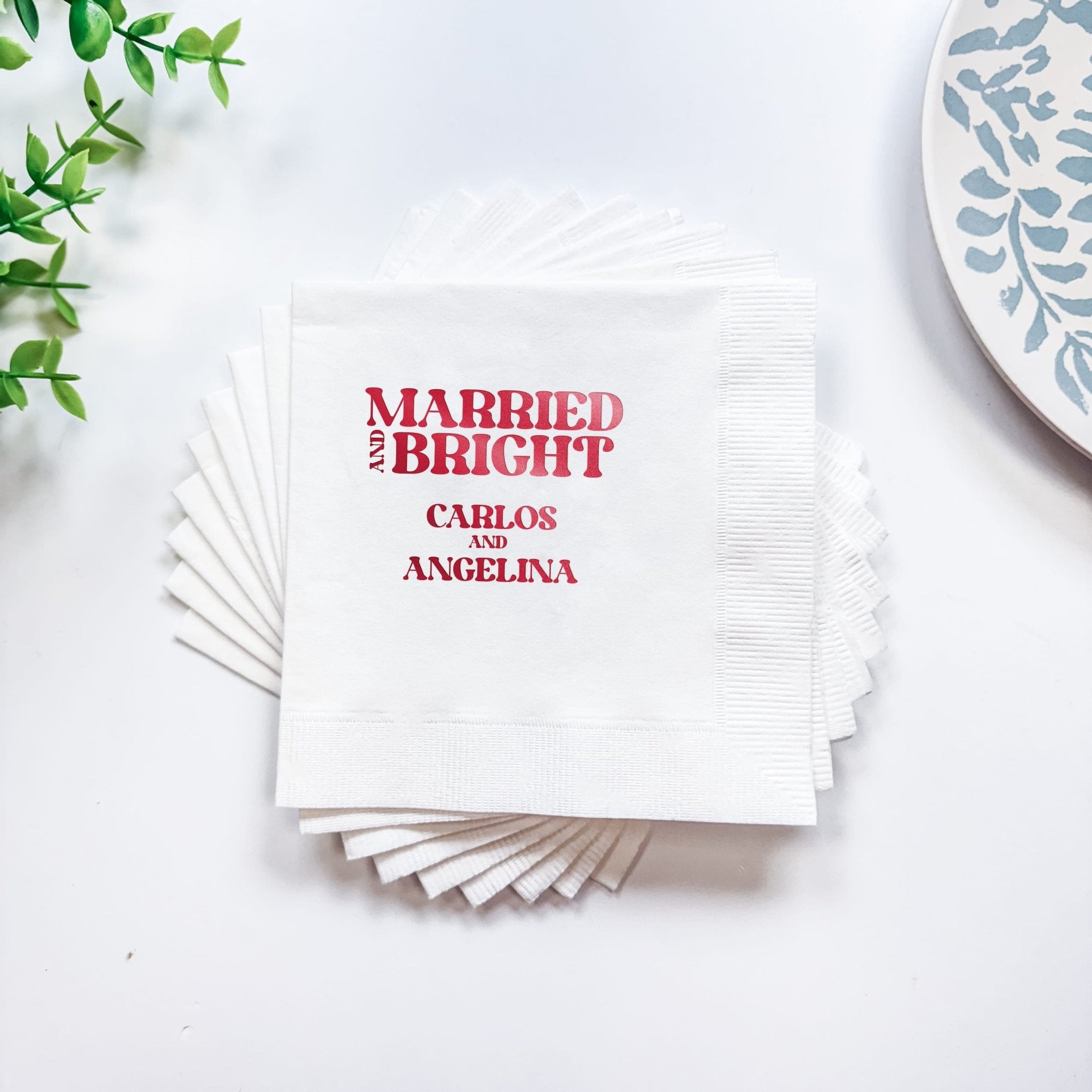 Stack of white cocktail napkins with red foil-stamped text reading "Married & Bright" and personalized names Carlos and Angelina