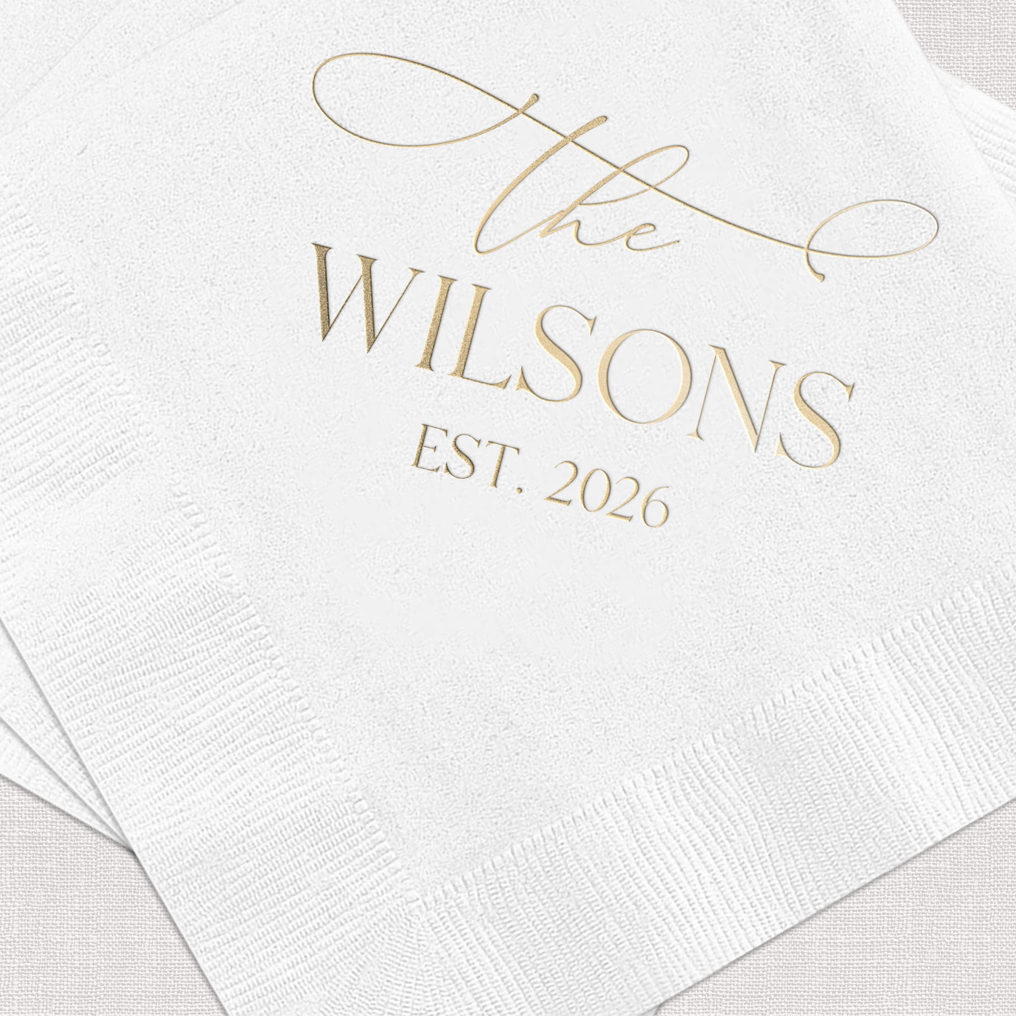 White cocktail napkins with gold foil stamped design reading "the Wilsons EST. 2026" with decorative flourishes