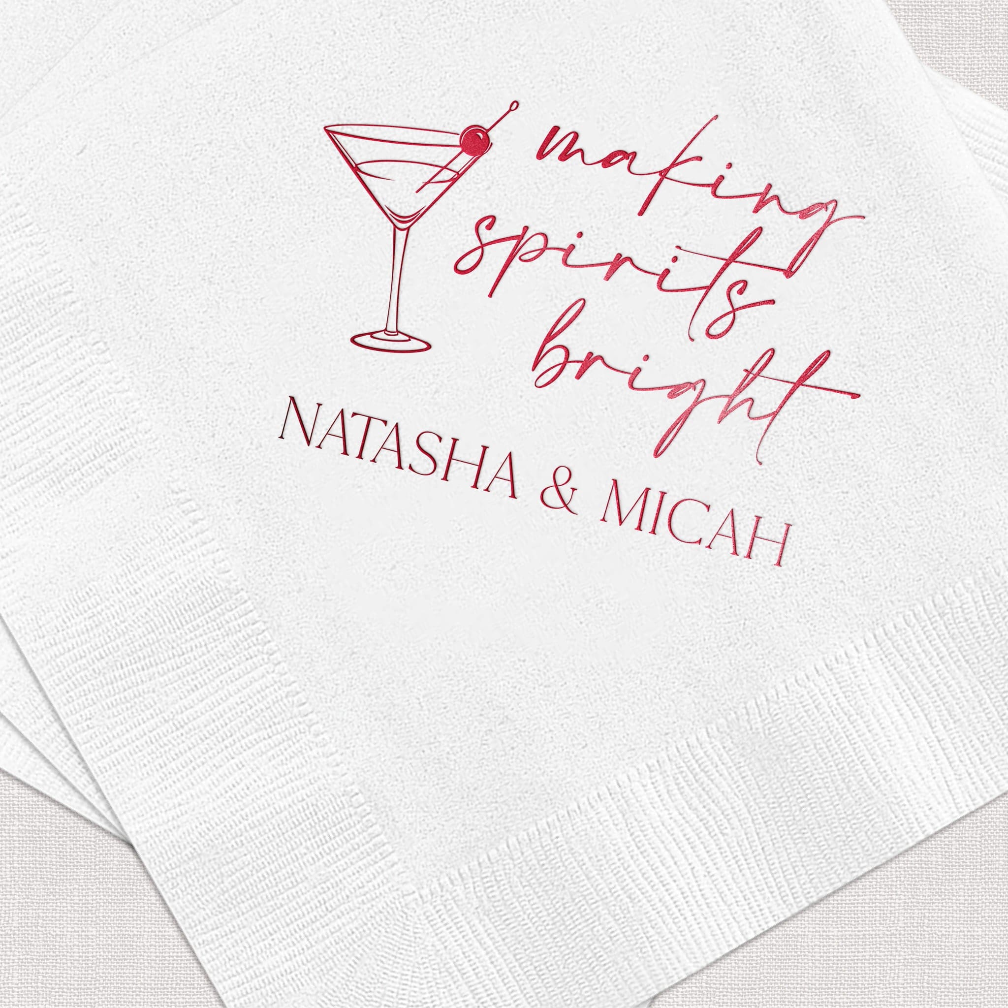 White beverage napkin with pink foil-stamped martini glass and "making spirits bright" text, personalized with "NATASHA & MICAH" below.