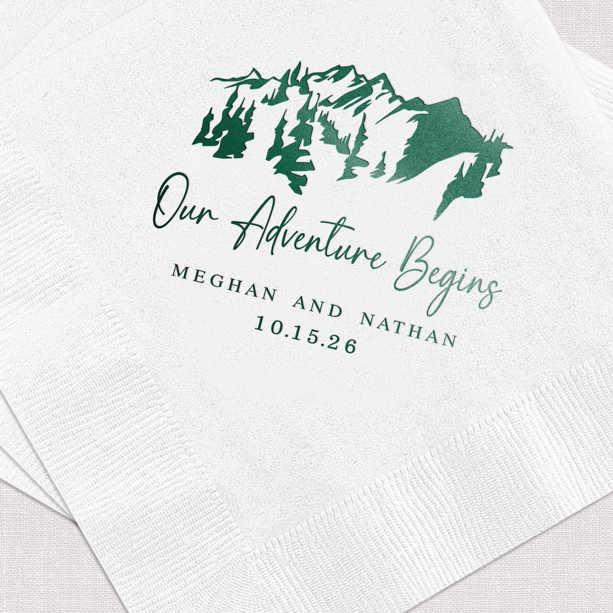 White napkin with green foil-stamped mountain design and "Our Adventure Begins" text personalized with names and wedding date.