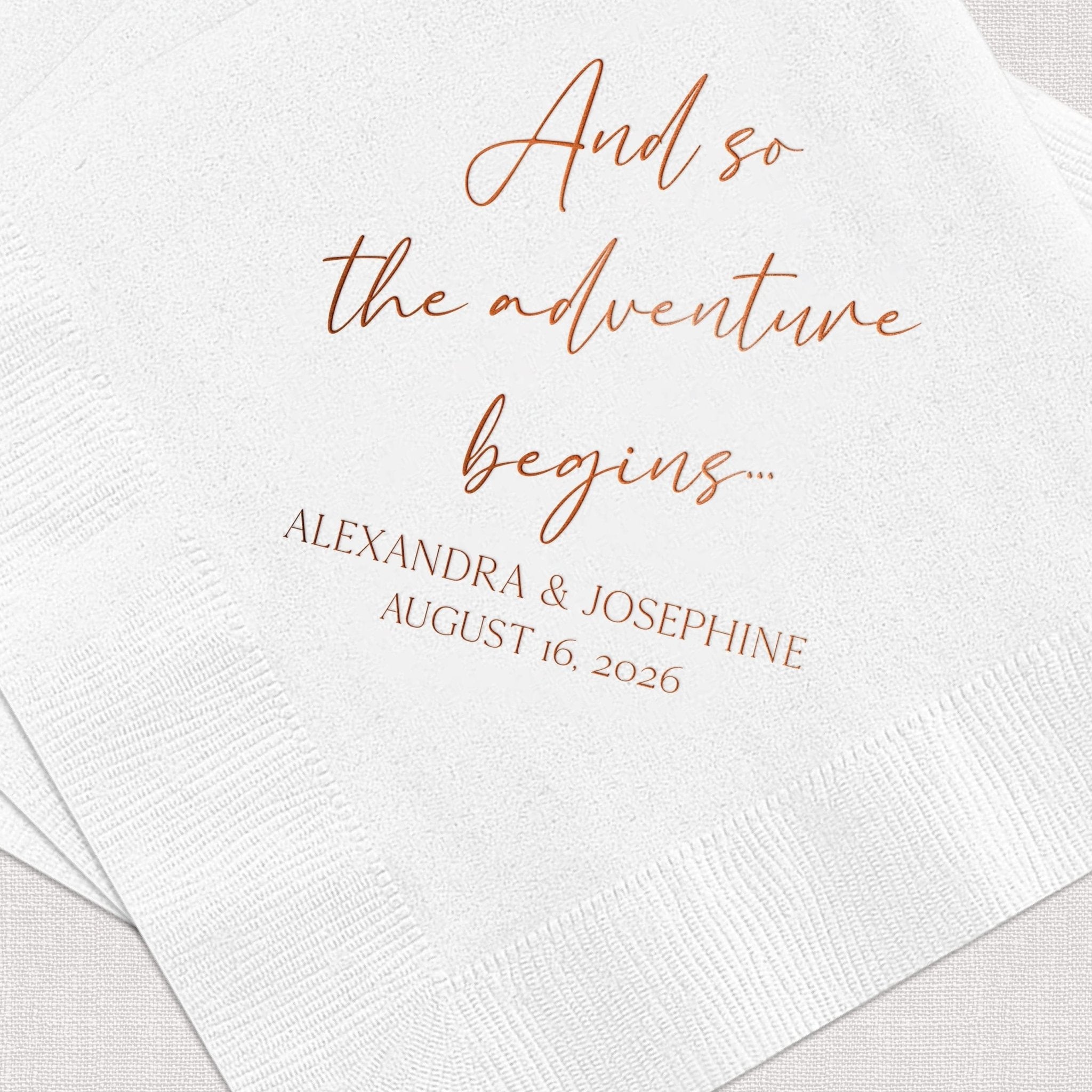 White cocktail napkin with rose gold foil text reading "And so the adventure begins" and personalized names "Alexandra & Josephine" with wedding date August 16, 2026.