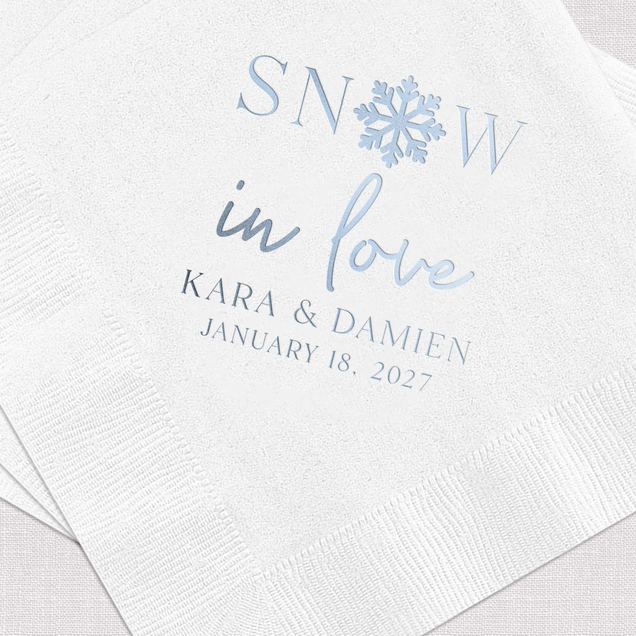 White beverage napkins with blue foil-stamped "Snow in Love" text, snowflake design, and personalized names Kara & Damien with wedding date January 18, 2027.