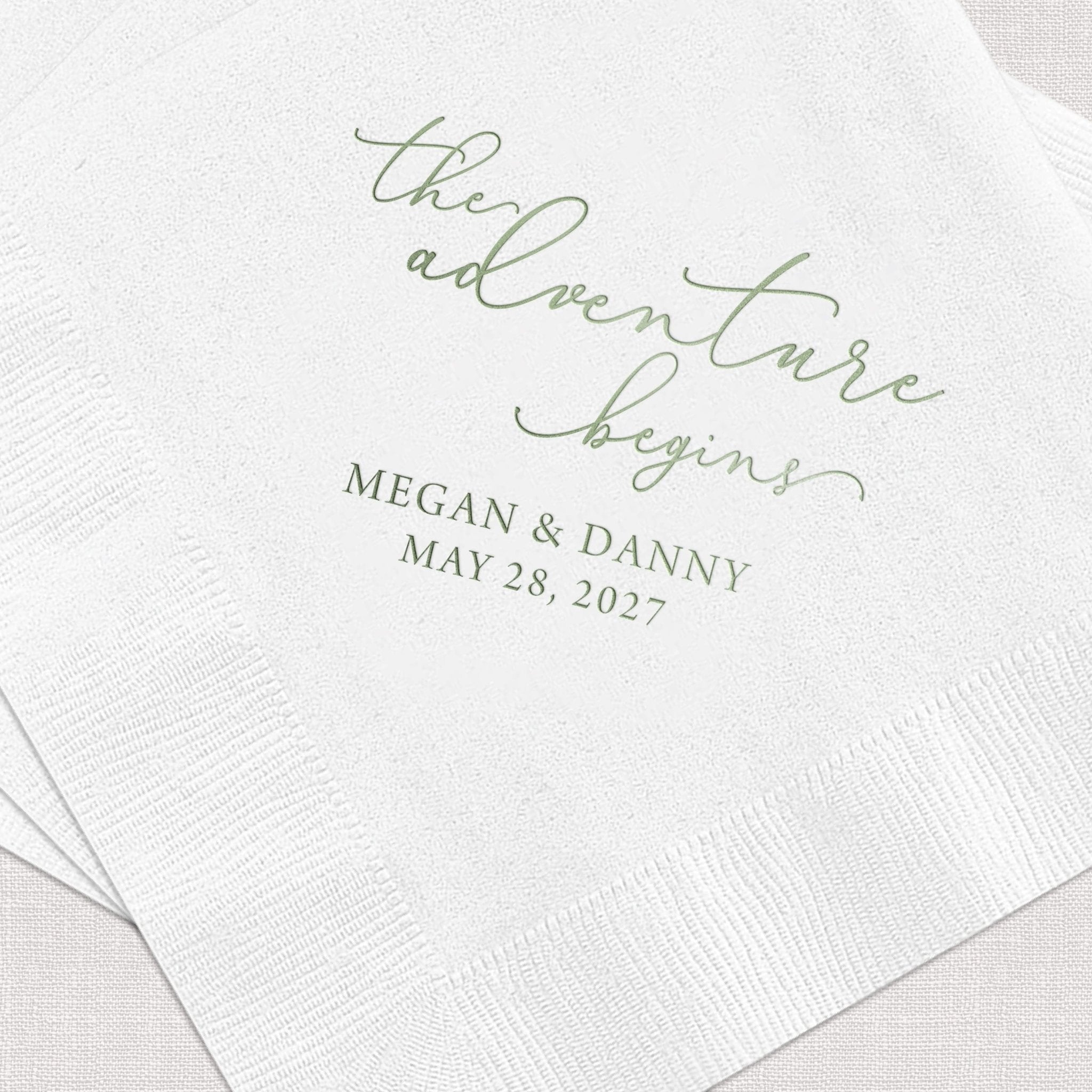 White cocktail napkins with sage green foil stamped text reading "The Adventure Begins" and "Megan & Danny May 28, 2027"