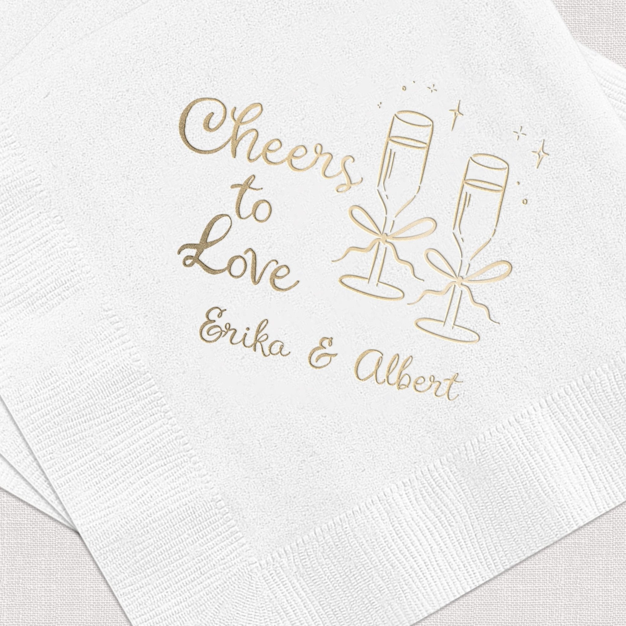 White cocktail napkin with gold foil stamped design featuring "Cheers to Love" text, champagne glasses, and personalized names Erika and Albert