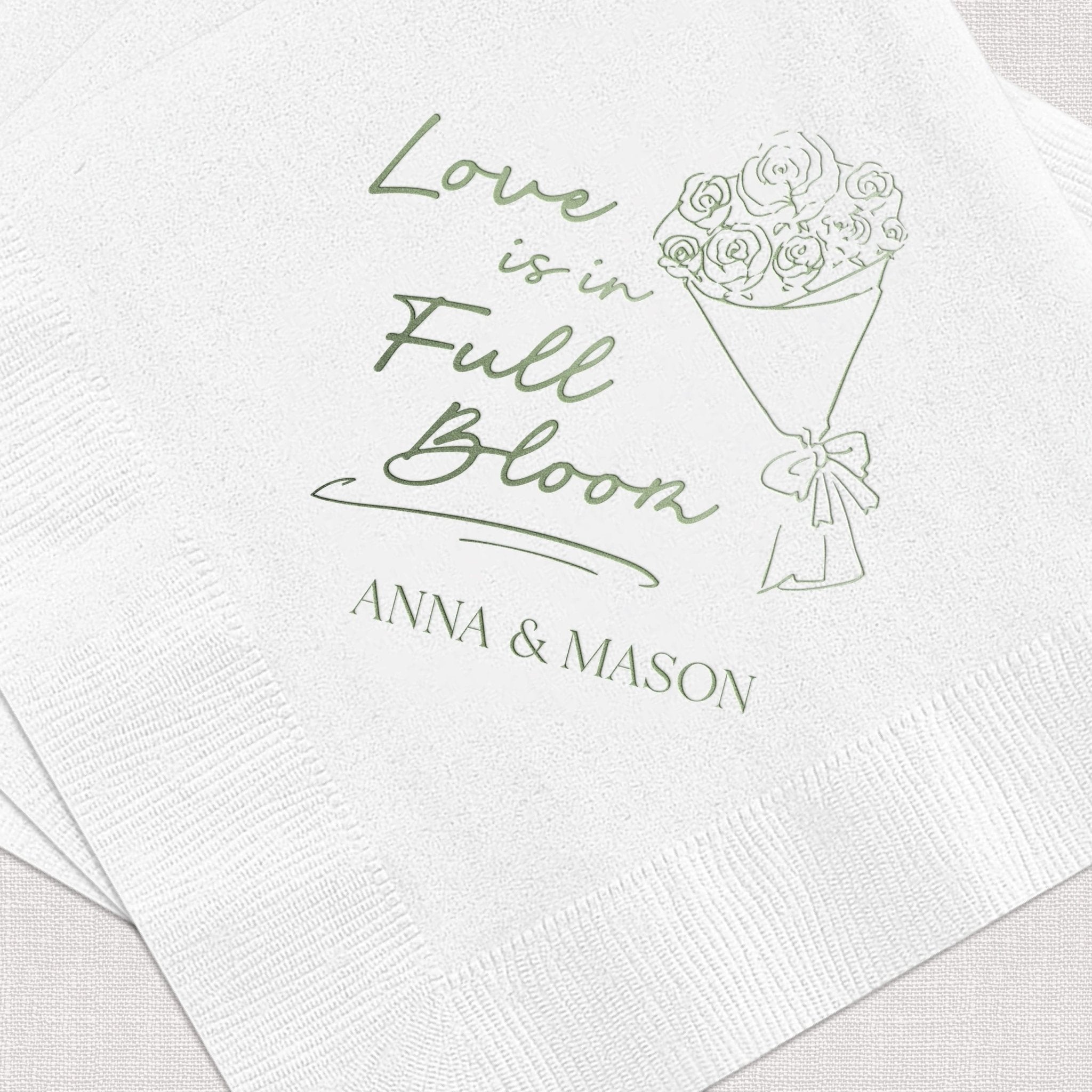 White napkin with sage green foil-stamped design featuring "Love is in Full Bloom" text, floral bouquet illustration, and personalized names Anna & Mason
