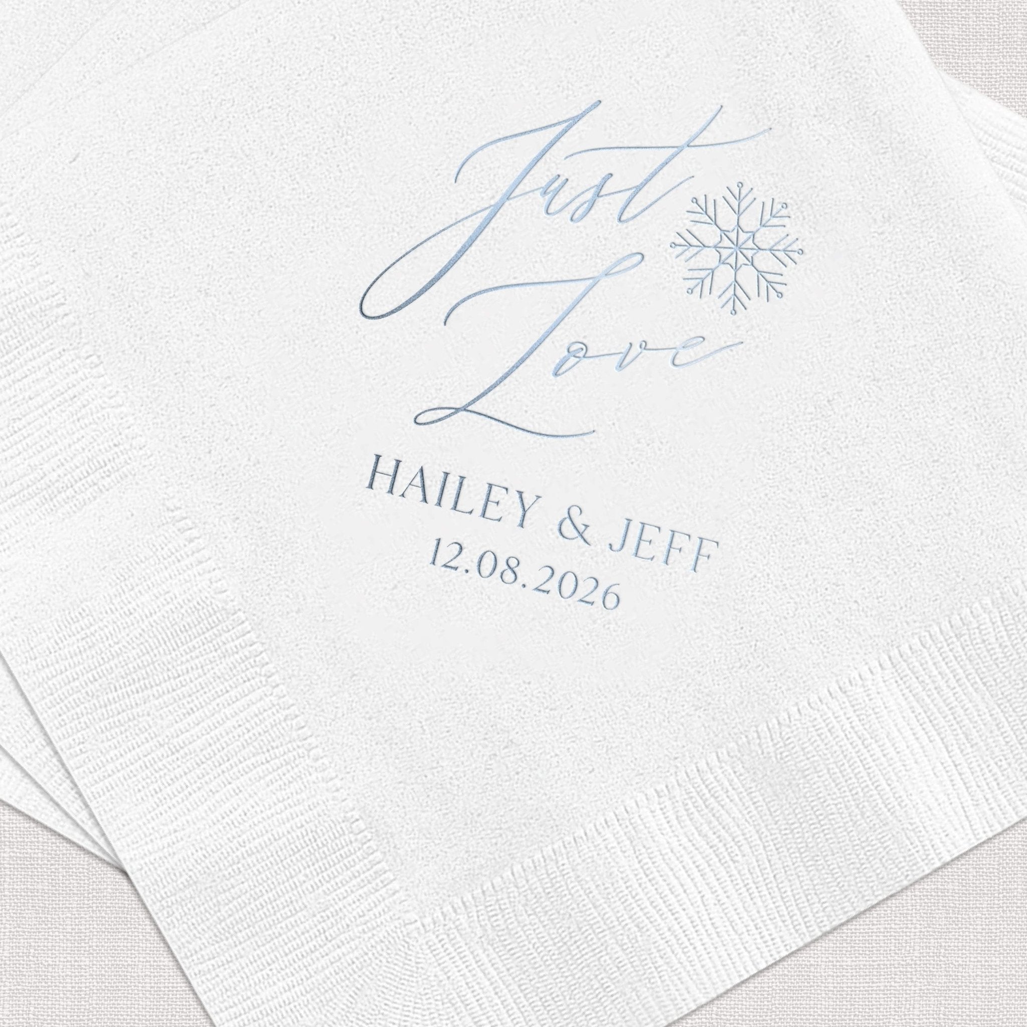 White cocktail napkins with blue foil-stamped "Just Love" text, snowflake design, and couple names Hailey & Jeff with wedding date 12.08.2026