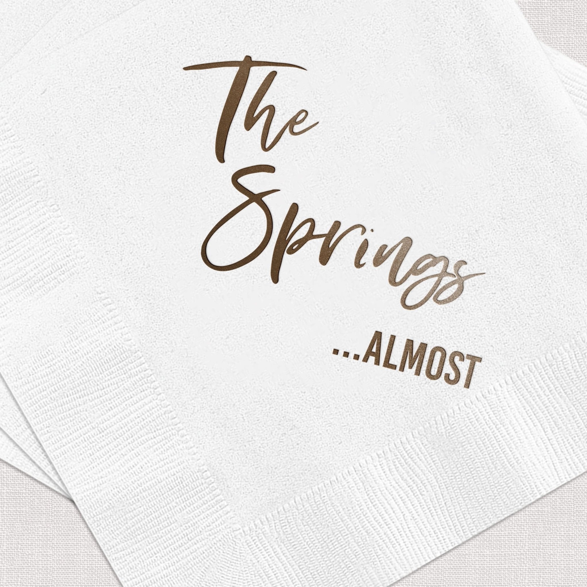 White cocktail napkins with "The Springs ...ALMOST" in gold foil lettering for bachelor party or wedding favors.