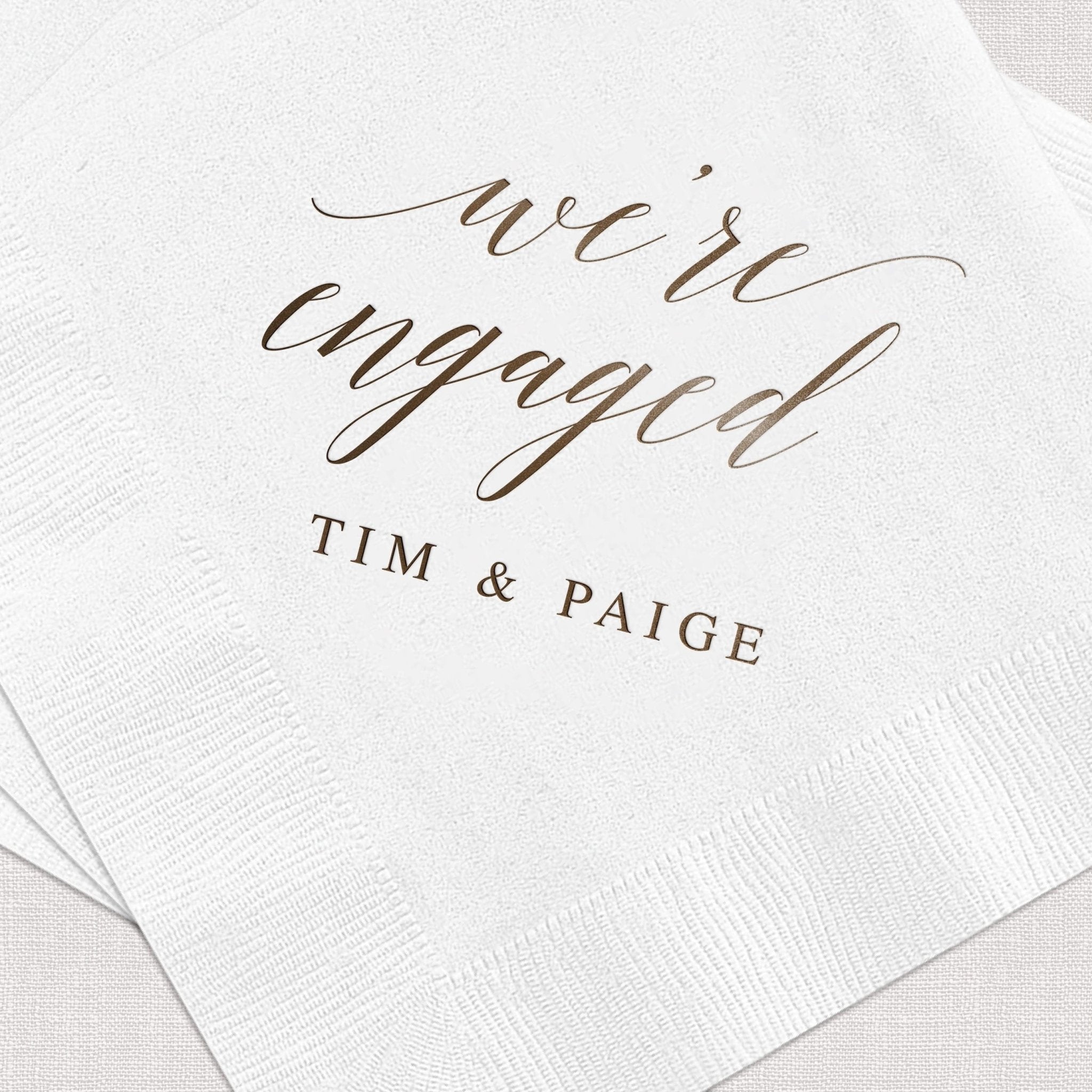 White cocktail napkins with gold foil stamped "we're engaged" script and "TIM & PAIGE" personalization text