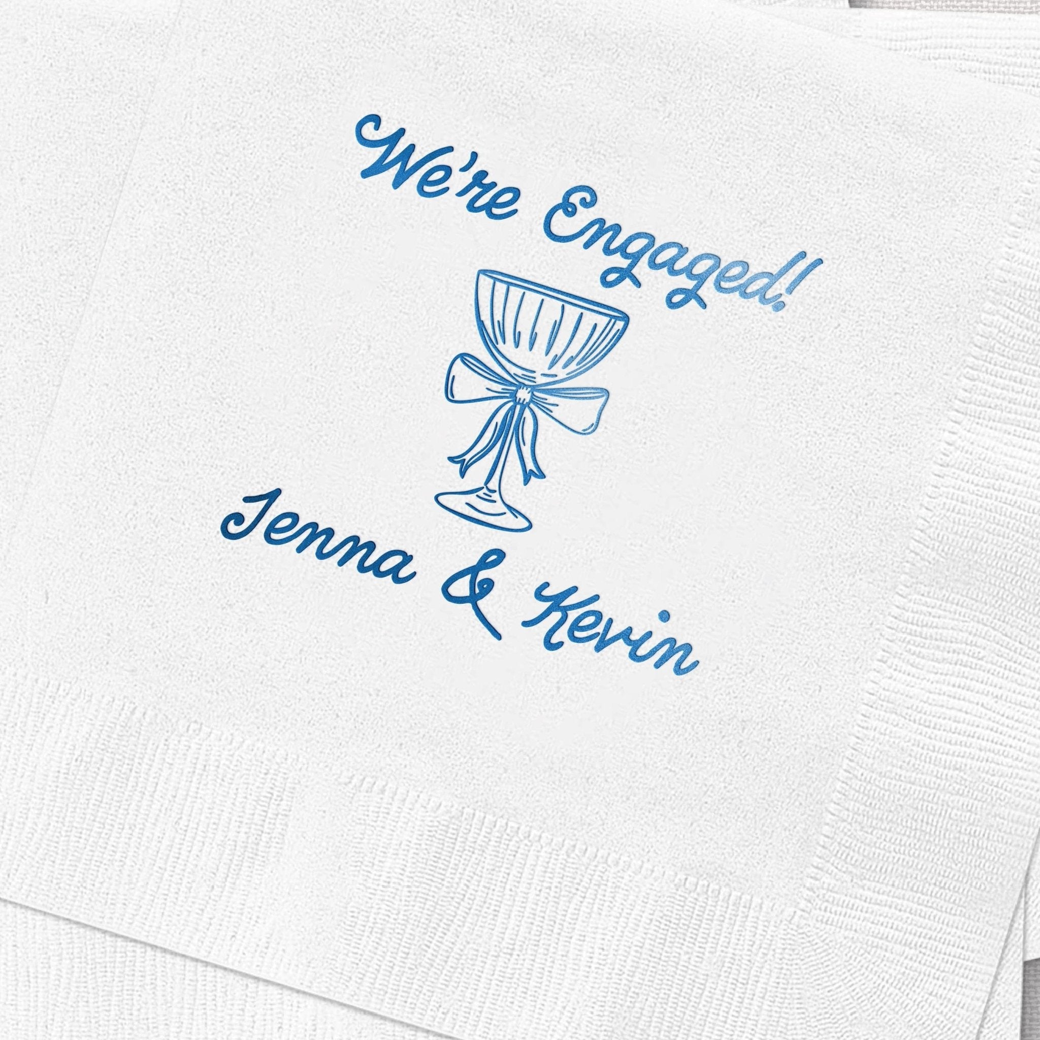 White cocktail napkin with blue foil-stamped design reading "We're Engaged!" above a bouquet illustration and personalized names "Jenna & Kevin" below.
