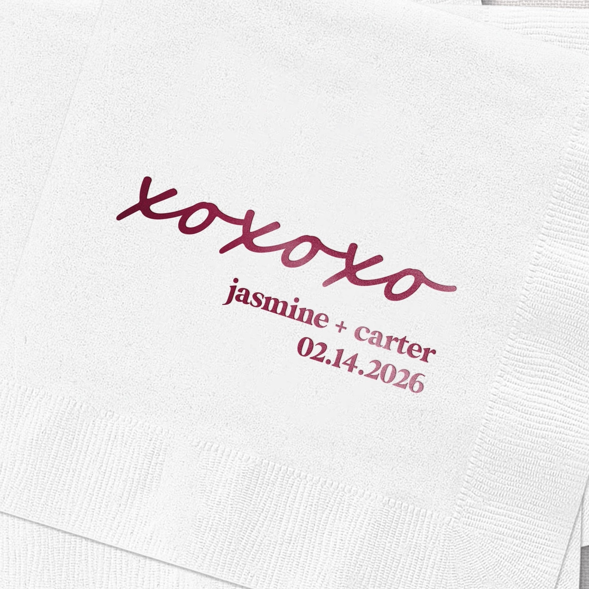 White cocktail napkin with burgundy foil-stamped XOXO design and personalized names "jasmine + carter" with date "02.14.2026"