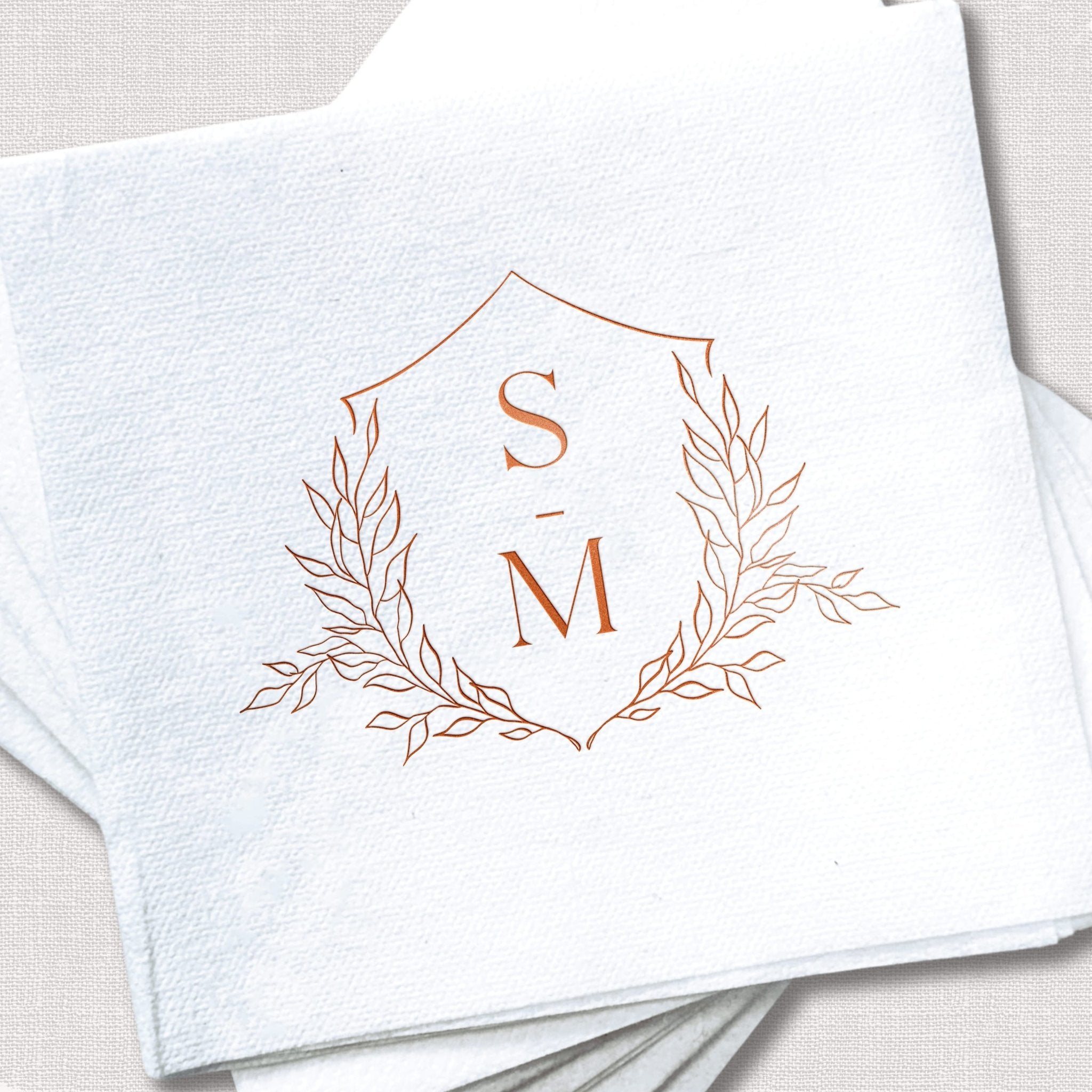 White linen napkin with rose gold foil-stamped floral crest design featuring initials S and M surrounded by delicate leaf branches