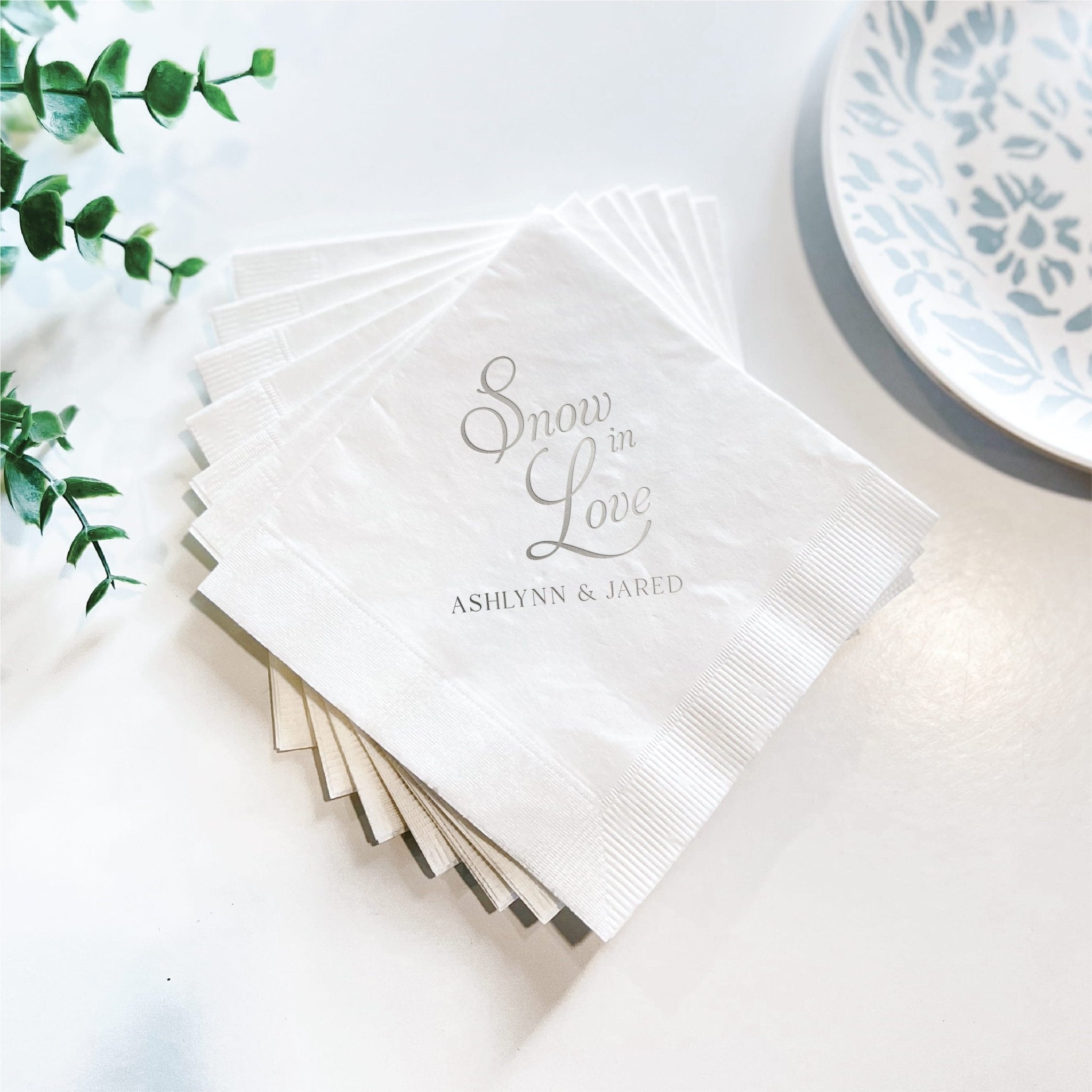 Stack of white napkins with "Snow in Love" and personalized names in gray foil stamping, arranged on white surface with greenery and decorative plate.