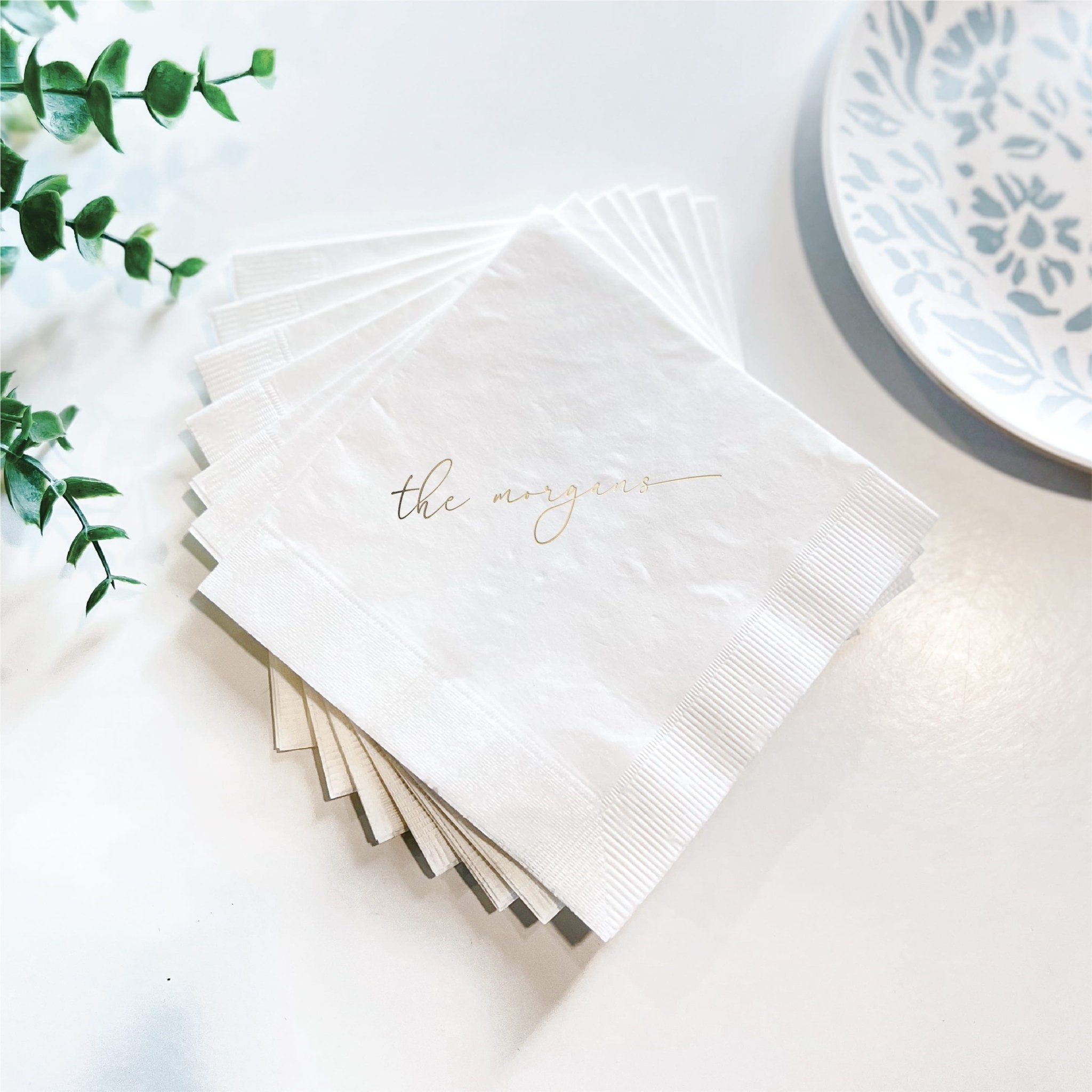 White cocktail napkins with foil-stamped last name, fanned stack beside blue and white plate and greenery