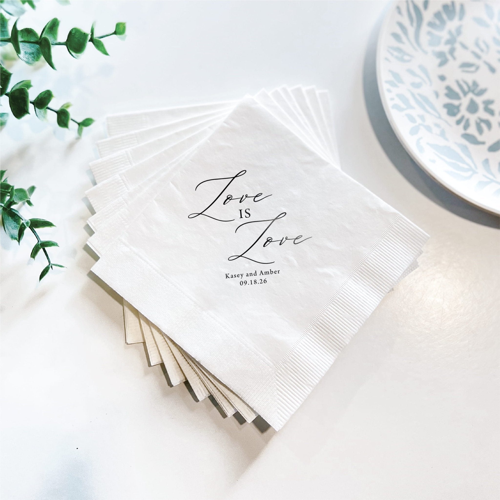 Stack of white napkins with "Love is Love" script and personalized names and date printed in black, styled with greenery and blue patterned plate.