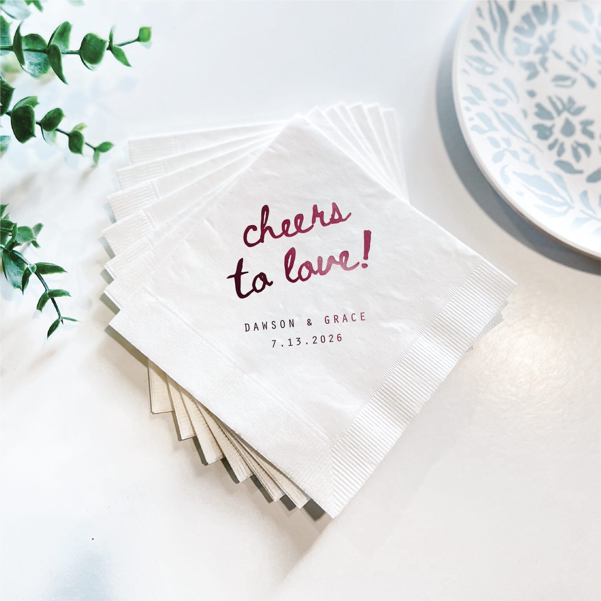 Stack of white 3-ply napkins with burgundy foil "cheers to love!" text and personalized names and date below, displayed with greenery and blue patterned plate.