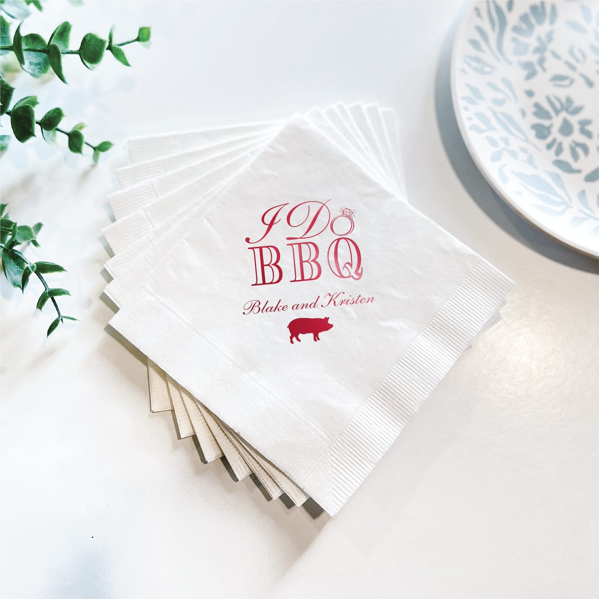 Stack of white napkins with pink foil-stamped "I Do BBQ" text, couple's names, and small pig icon for wedding party.