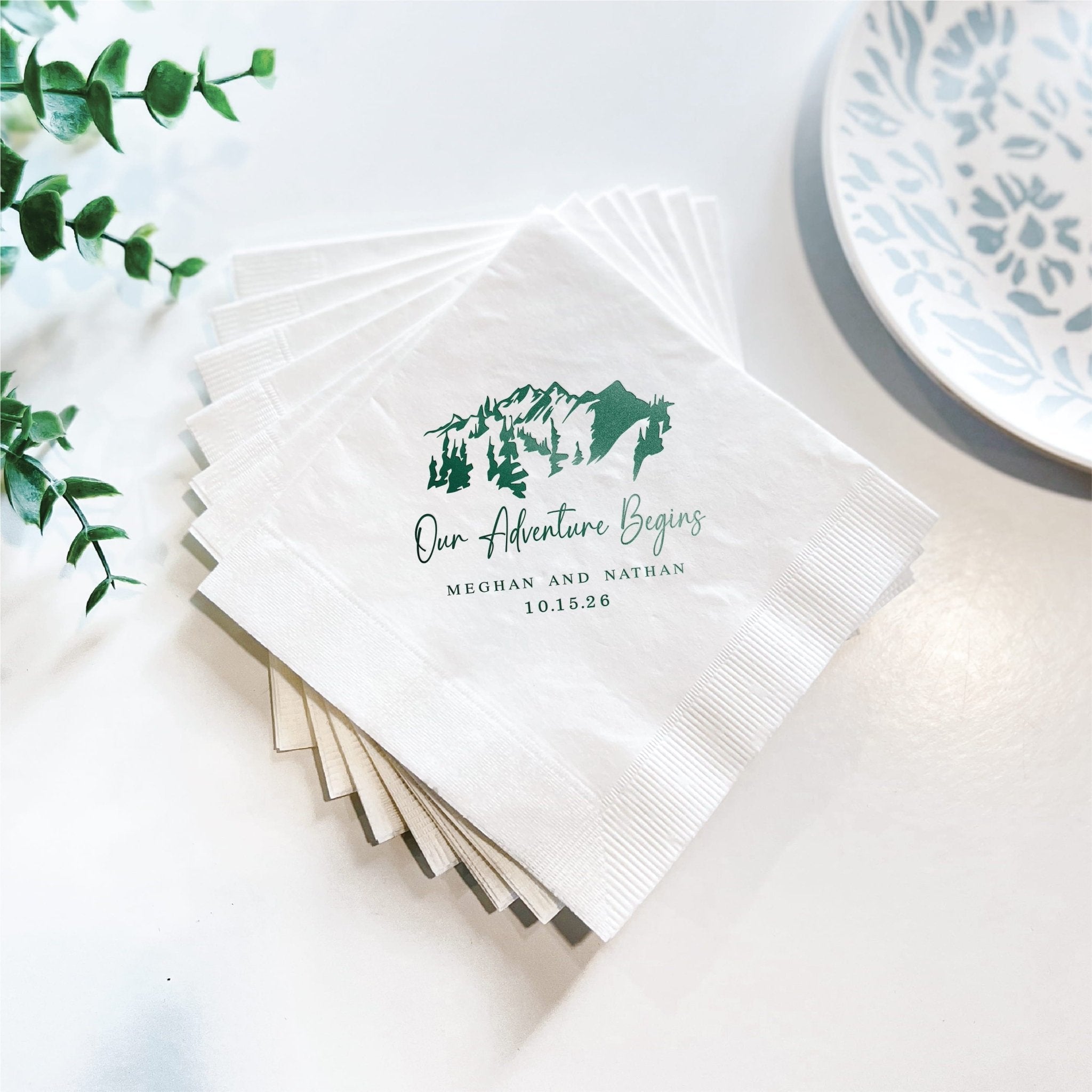 Stack of white napkins with green foil-stamped mountain design and "Our Adventure Begins" text with couple names personalized below.