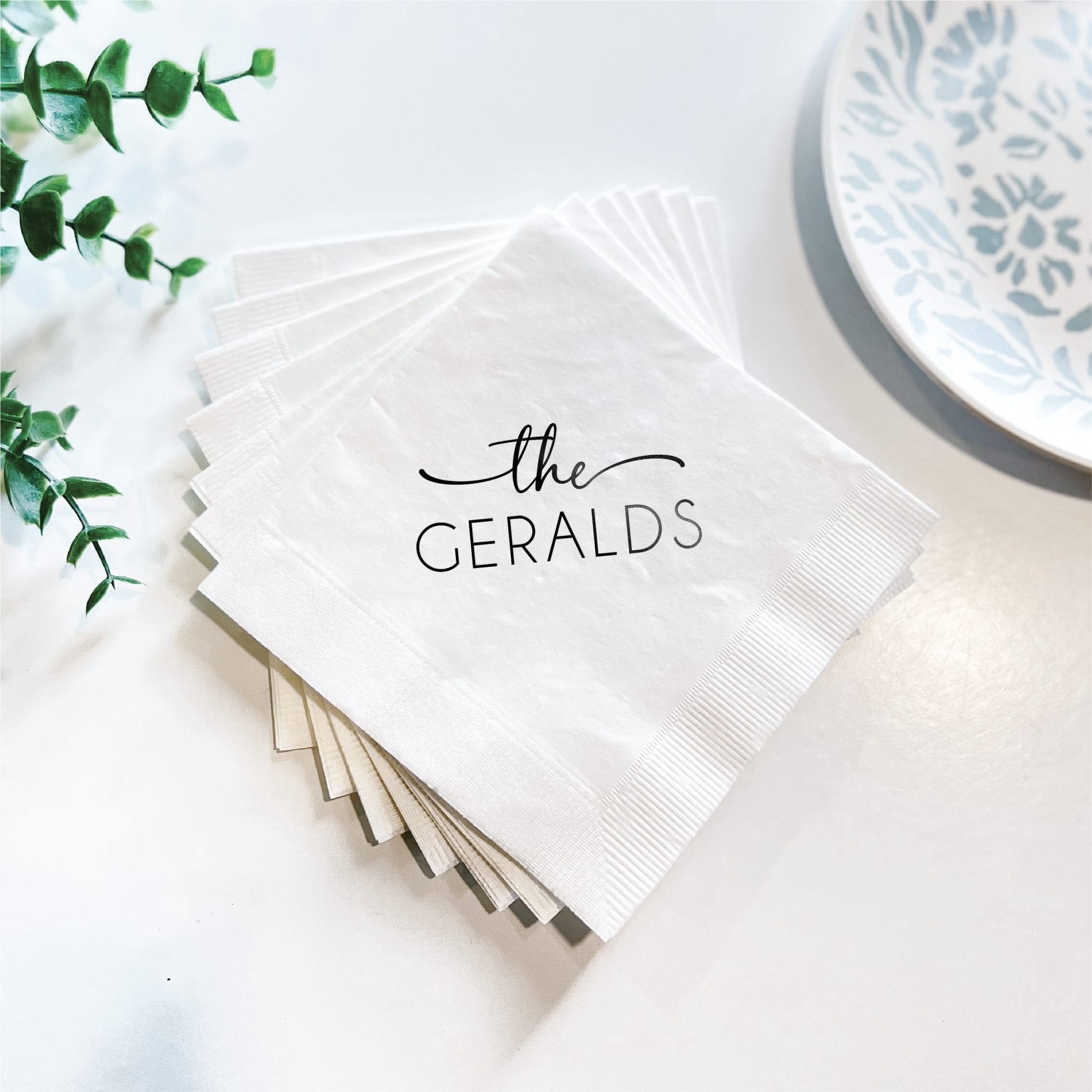Stack of white personalized napkins with "the GERALDS" printed in black text, displayed with greenery and blue patterned plate.